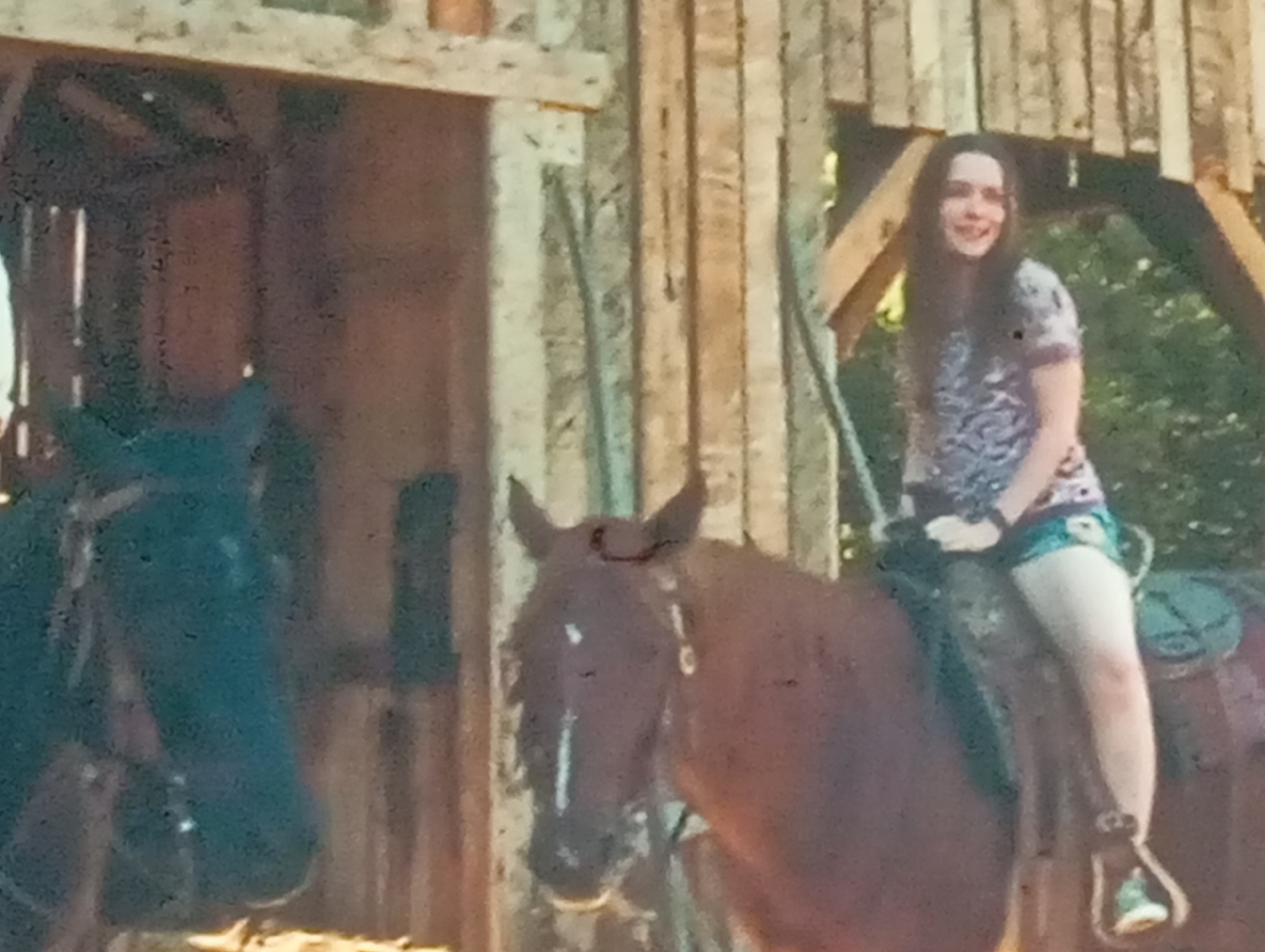 The media file depicts a young woman riding a horse in a rustic setting, possibly a stable or barn. She is seated on a brown horse, with a darker horse visible to her left. The woman has long, dark hair and is smiling. She is wearing a patterned t-shirt and shorts. The background features wooden structures, suggesting a rural or agricultural environment. The overall impression is one of a casual outdoor activity. The lighting suggests it might be daytime, with natural light illuminating the scene. There are no visible signs of text in the image. The context provided places the scene in Sofiivska Borshchahivka, Ukraine.