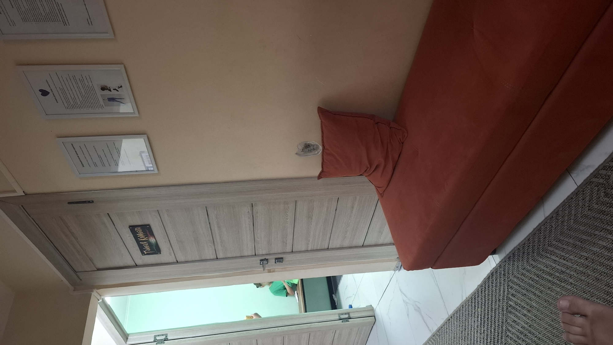 A low-angle, eye-level shot depicts an interior scene, likely a waiting area or treatment room. The foreground is dominated by a foot and a portion of a textured rug on the left. To the right, an orange upholstered bench with a matching pillow rests against a peach-colored wall. The wall features a distressed area above the bench and two framed documents to the left, one with a heart symbol and the other with text. A wooden door with horizontal paneling stands to the left of the bench. Through an open doorway on the far left, a person in a green shirt is visible in a room with a bright, greenish-blue light and a wooden bench. The scene suggests a professional establishment, possibly a spa or clinic. No specific location in Shymkent, Kazakhstan, can be identified from the visual evidence.