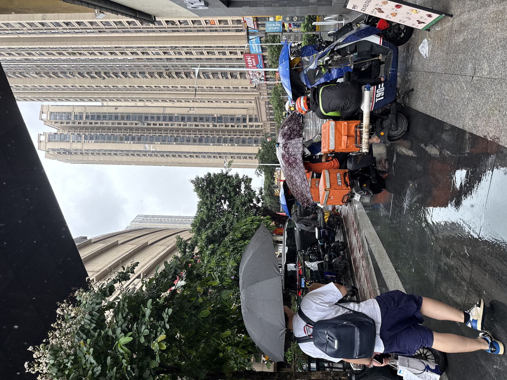 On a rainy day in Haizhu District, China, a person walks with an umbrella towards the viewer, carrying a backpack and shopping bags. In the background, several delivery workers are gathered with their motorcycles, which are equipped with orange insulated delivery boxes. Tall residential buildings dominate the skyline. The street is wet and reflective due to the rain, and the sky is overcast.