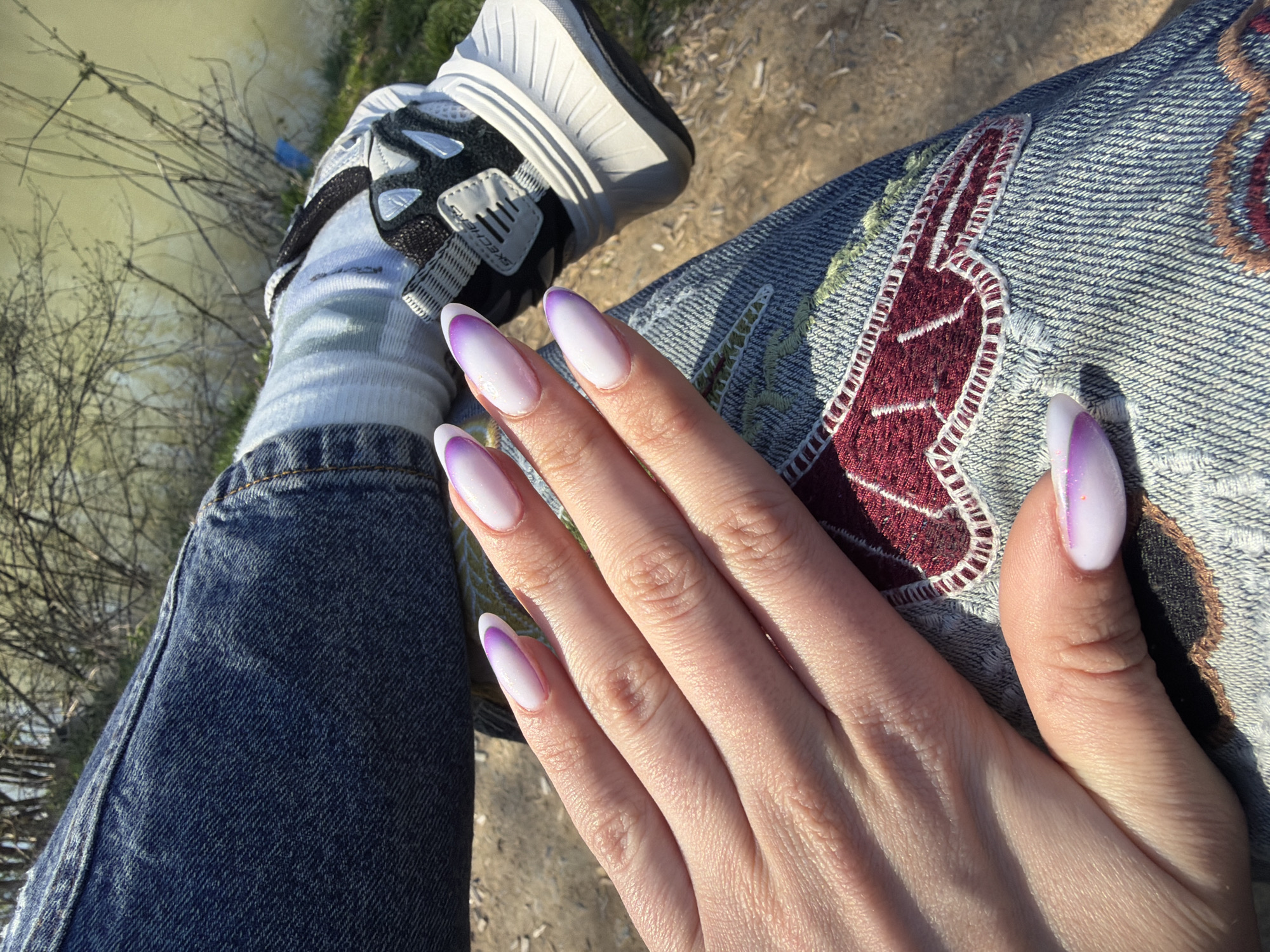 The image shows a person sitting outdoors, likely near a body of water. The person is wearing blue jeans with an embroidered design and white socks with black and white sneakers. The focus is on the person's hand, which is resting on their leg. The nails are painted with a white and purple ombre design. The background includes a glimpse of water, possibly a river or lake, and some vegetation. The image does not contain enough details to identify the exact location in Stara Zagora, Bulgaria.