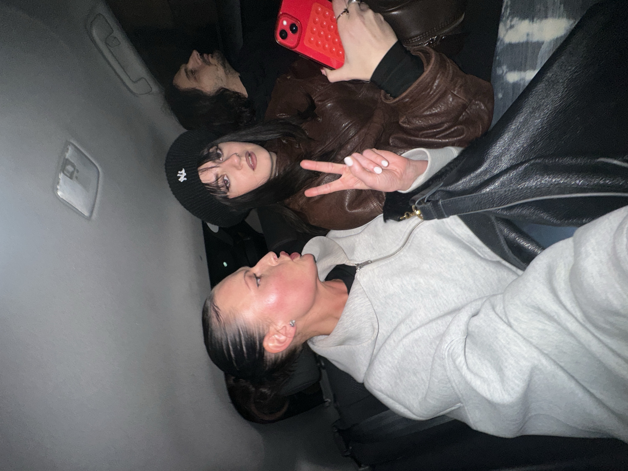 The image depicts three individuals seated in the rear passenger compartment of a private vehicle at night, within the city of London, United Kingdom.
On the left, a woman with dark hair pulled back into a sleek ponytail wears a light grey zippered hoodie. Her head is turned sharply to the right, with her mouth slightly open. Her cheeks exhibit a distinct pink flush, and she wears a small stud earring.
In the center, a second woman with dark hair and bangs wears a black beanie with a white "NY" logo and a brown leather jacket over a black long-sleeved top. She holds a red smartphone, fitted with a red, textured grip accessory, in her left hand. Her right hand is raised, forming a peace sign with white-painted fingernails. She has a nose piercing and gazes forward.
Partially visible in the right background is a man with long, dark, curly hair and a beard, wearing a dark top. He is observed in profile, looking towards the right side of the frame.
The vehicle's interior is dimly lit, featuring a grey headliner and a rectangular overhead light fixture. A black shoulder bag with a gold clasp is visible in the lower-left foreground.