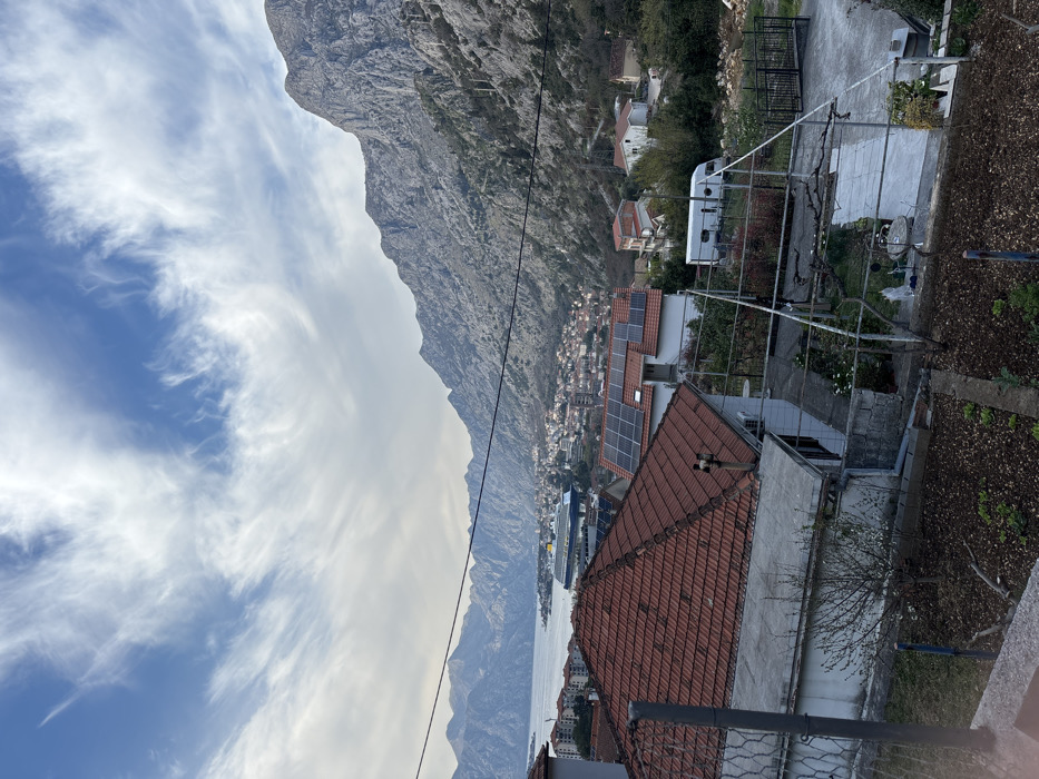 An elevated panoramic view captures the city of Škaljari, Montenegro, overlooking the Bay of Kotor and its surrounding mountains. The sky features a mix of blue patches and scattered, wispy white clouds. Rugged, grey limestone mountains dominate the background, particularly on the right side, with sparse green vegetation on their lower slopes.
In the mid-ground, the calm waters of the Bay of Kotor are visible, where a large blue and yellow cruise ship is moored or anchored. The shores of the bay are lined with numerous residential buildings, characterized by red-tiled roofs, forming a dense urban landscape that extends up the lower mountain slopes.
In the immediate foreground, a multi-tiered residential area is visible. The closest structure on the left features a flat grey roof section and a red-tiled roof with a chimney. Adjacent to it, another house prominently displays multiple dark solar panels on its red-tiled roof. To the right of these houses, a white recreational vehicle (RV) is parked in a fenced yard, bordered by a black metal gate further right. The immediate lower foreground consists of a garden plot with tilled soil and small green plants, supported by a basic trellis structure and enclosed by low concrete walls and a partial metal fence. Utility lines traverse horizontally across the mid-ground. No human or animal figures are present in the scene.