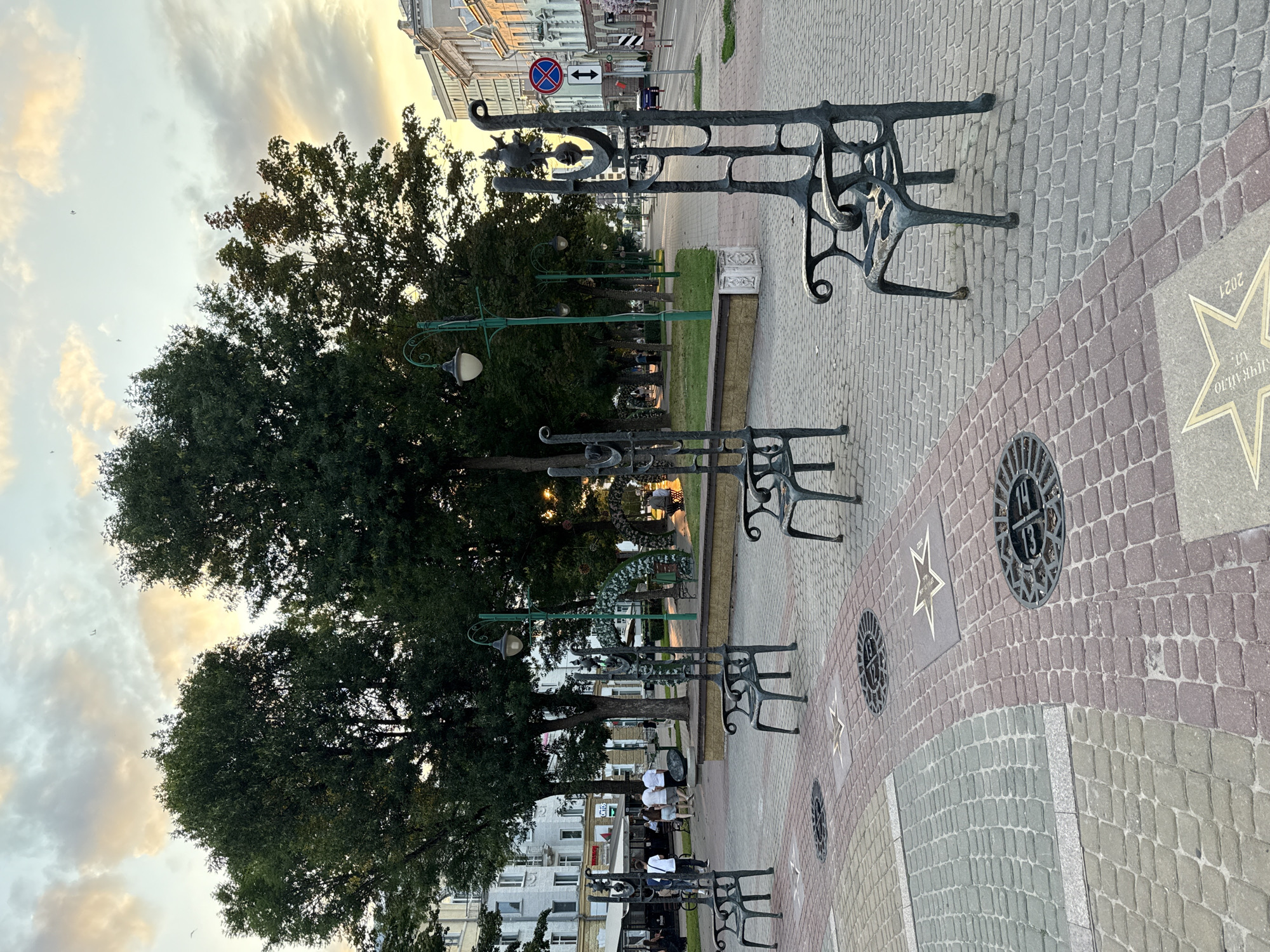 This image depicts a street scene in Mahilyow, Belarus, with a focus on a paved pedestrian area featuring several ornate, dark metal chairs. The chairs are sculptural and have a distinctive design, appearing to be made of cast metal. They are placed at intervals on a patterned brick walkway. Some of the bricks are a reddish-brown, while others are a greyish-green. Inlaid into the walkway are circular metal grates and several star-shaped plaques, some of which appear to have inscriptions.
In the background, there are mature trees lining a park-like area. Beyond the trees, buildings are visible, including a multi-story structure with windows and awnings, and further down the street, a more modern building. Streetlights with a green design are present along the walkway.
Several people are walking in the distance, appearing as small figures in the mid-ground. One group of four individuals, two men and two women, are walking together on the sidewalk. A car is visible on the street in the far background, and a traffic sign with a red circle and a blue background is also present. The sky is overcast with clouds, and the lighting suggests it is either early morning or late afternoon. The overall impression is of a public urban space designed for relaxation and possibly commemoration, given the star plaques.