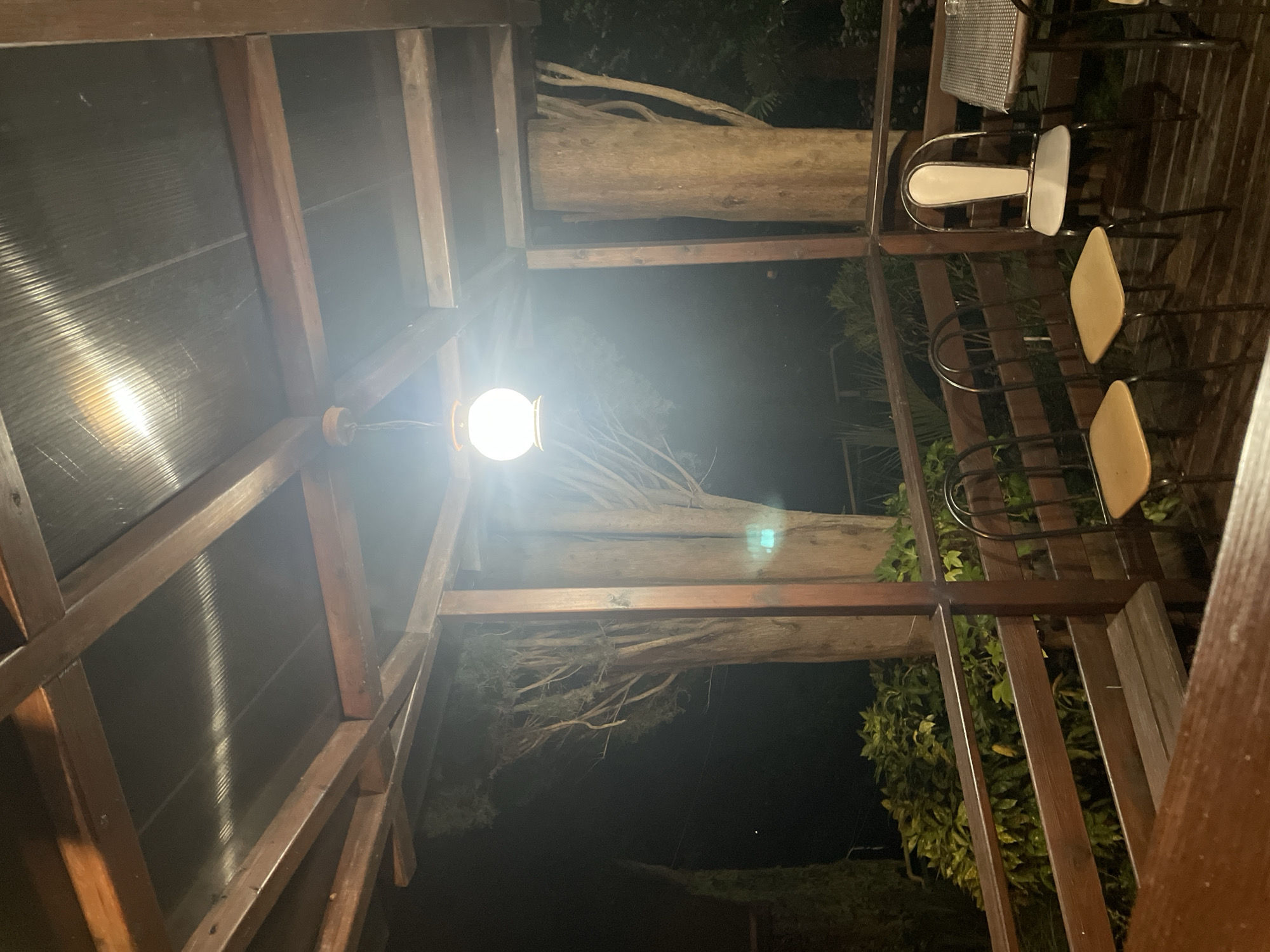 The image shows an outdoor dining area at night, likely on a balcony or porch. A wooden structure with a translucent roof is visible overhead, supported by wooden beams. A single, glowing lantern hangs from the ceiling, illuminating the scene. In the background, large trees with rough bark are silhouetted against the dark night sky. A wooden railing encloses the area, and several chairs are arranged around small tables. The ground is made of wooden planks. A wooden bench is also present. The overall atmosphere is dimly lit and appears to be a quiet, outdoor setting.