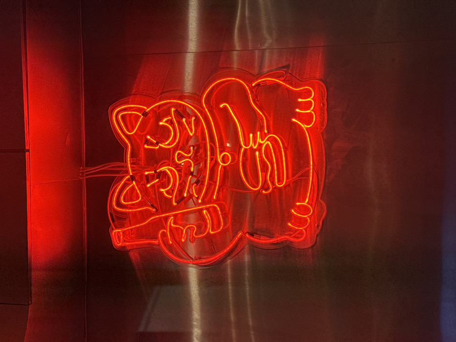 This image captures a brightly illuminated red neon sign depicting a stylized, cartoonish cat, resembling a Maneki-neko, against a brushed metallic wall. The cat figure features prominent ears, wide eyes, a small nose, and a simple mouth. Its raised left paw clutches a stylized bottle, while its right paw holds an object resembling a hamburger or bun. The intense red glow of the neon tubing vividly reflects off the textured metallic surface, creating intricate patterns of light and shadow. Visible wiring extends from the top of the neon sign. The surrounding environment appears dimly lit, enhancing the visual prominence of the red light. This scene is located in the city of Paris, France.