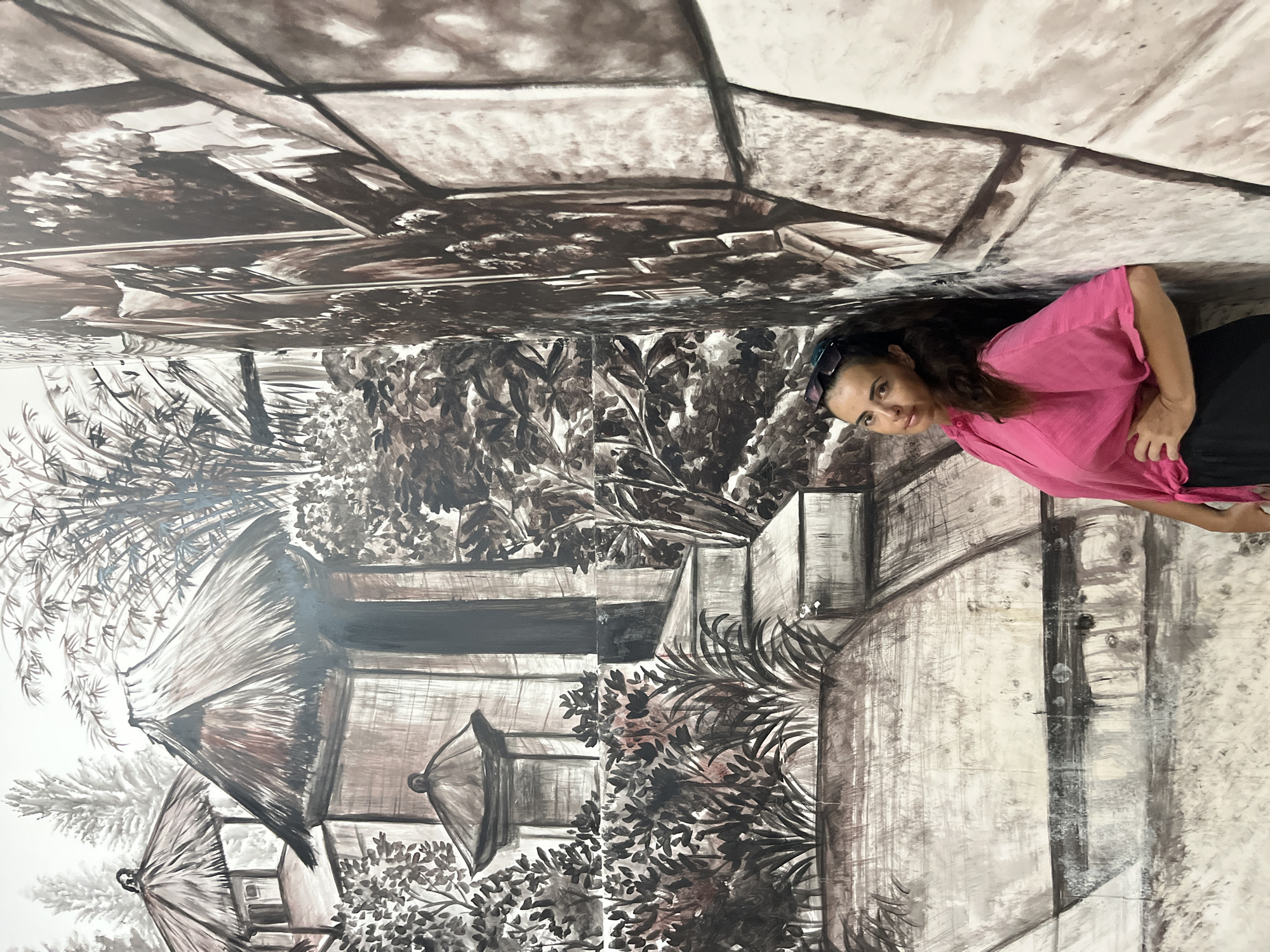 A medium shot captures a woman with dark, wavy hair and sunglasses perched on her head, leaning against a wall adorned with a monochromatic mural. She is positioned on the right side of the frame, her body angled slightly towards the viewer. She wears a vibrant pink, loose-fitting shirt tied at the waist and black pants. Her left hand rests on her hip, while her right arm is bent, with her forearm extending downwards.
The mural behind her depicts a stylized landscape with traditional Balinese architecture. On the left, structures with thatched roofs and stone elements are visible amidst lush foliage and bamboo stalks, rendered in shades of black, brown, and white. The right side of the mural transitions to a more abstract representation of textured walls and architectural details. The painting covers the entire wall, creating an immersive backdrop. The overall impression is that of a cultural or artistic installation, possibly located in a public space or gallery in Kuta Selatan, Indonesia. The lighting is even, suggesting an indoor setting or overcast conditions.