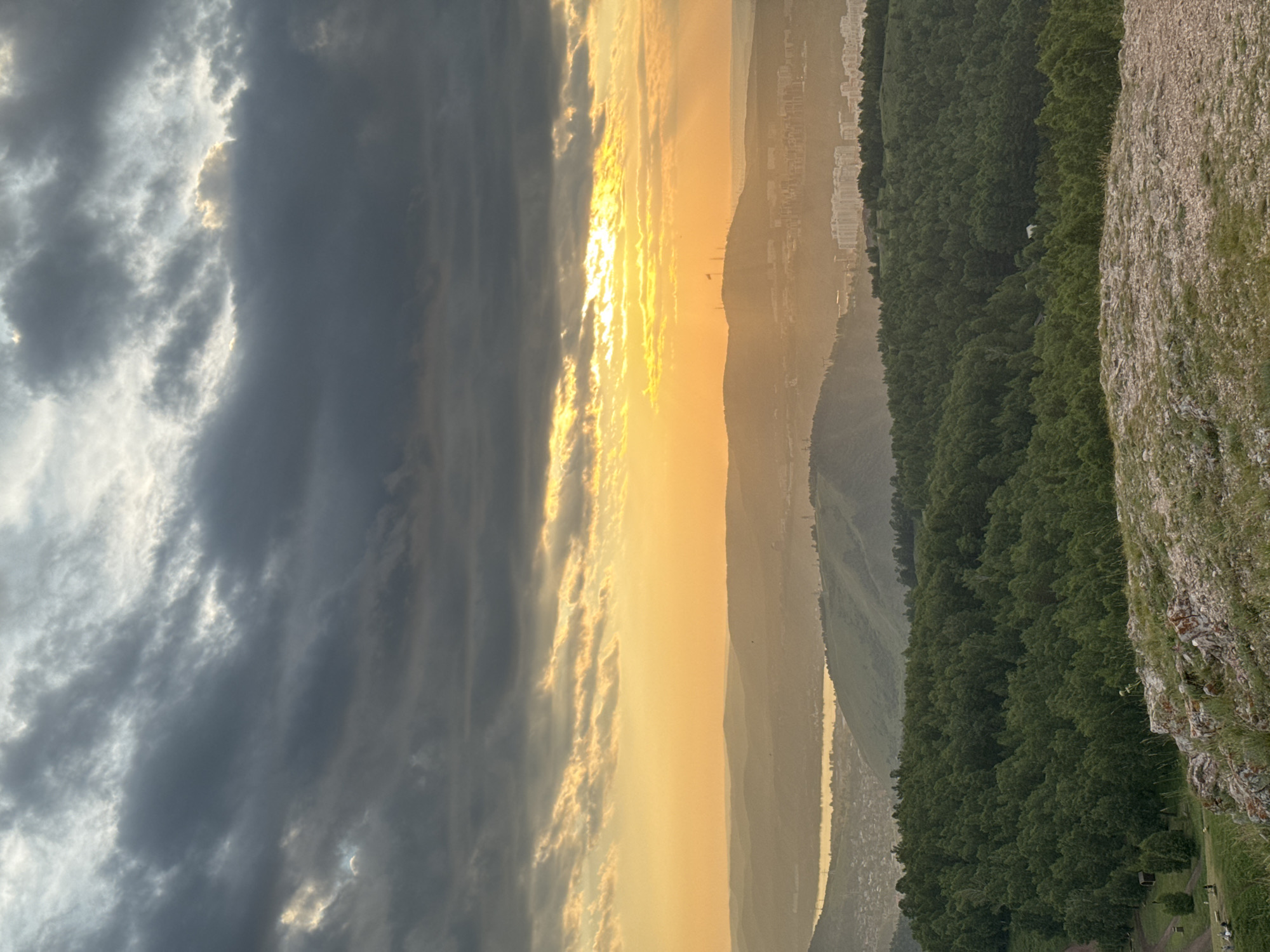 The image captures a panoramic view from a rocky, grassy hill overlooking the city of Krasnoyarsk, Russia, during sunset. The sky is dramatic, with dark, heavy clouds partially obscuring the setting sun, which casts a warm, golden-orange glow across the horizon. Rays of light pierce through the clouds, illuminating the layers of distant hills and the urban landscape.
In the foreground, the rocky terrain is covered with sparse green grass and a few wildflowers. A small wooden structure and some scattered people are visible on the grassy slope below.
Further in the distance, a dense, dark green forest blankets the rolling hills. Beyond the forest, the city of Krasnoyarsk unfolds with various buildings, including apartment complexes and a prominent hill topped with structures, possibly a monument or communication towers. A body of water, likely the Yenisei River, is visible to the left, reflecting the sunset light. The overall scene is one of natural beauty juxtaposed with an urban environment, under a striking twilight sky.