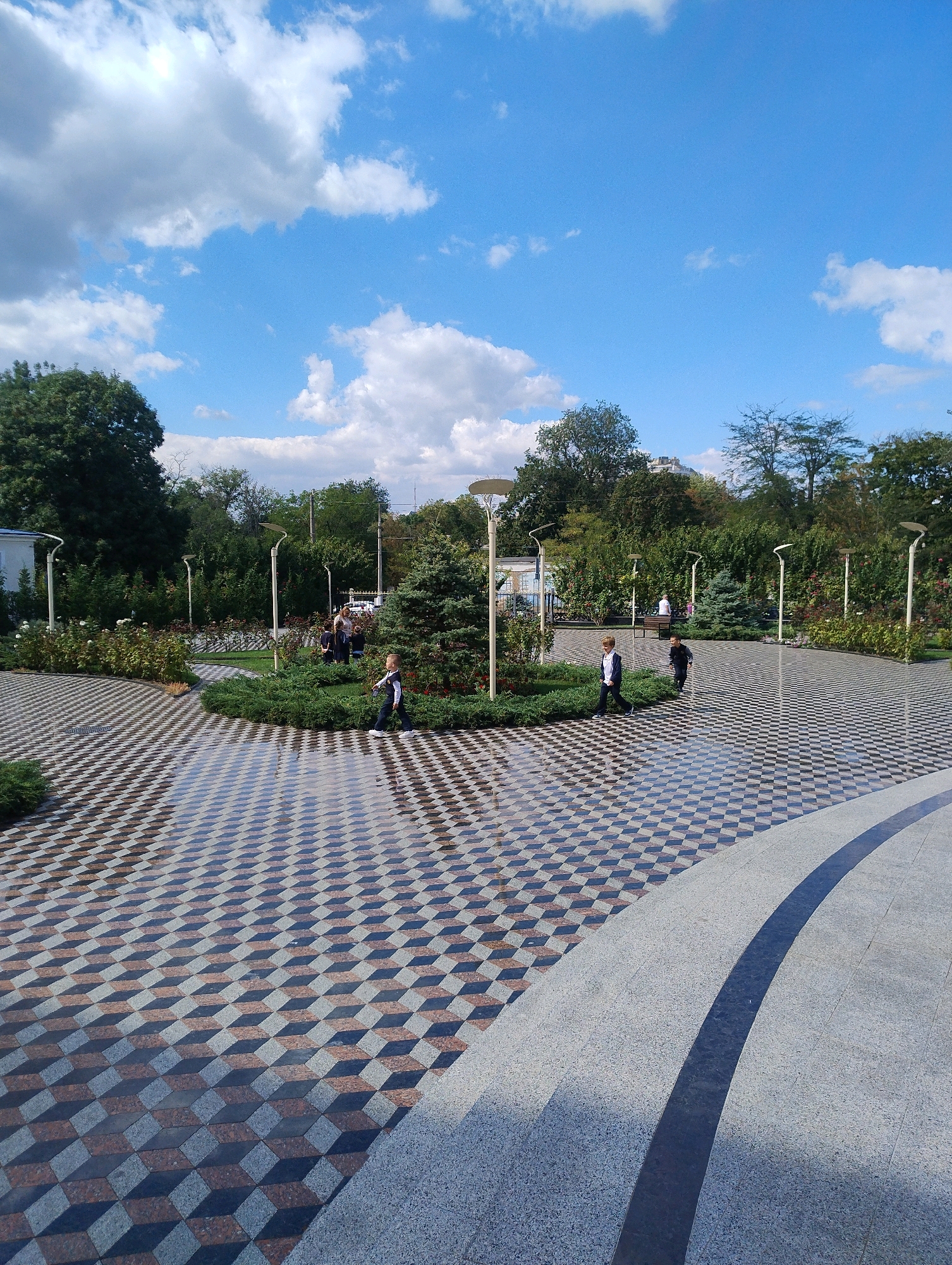 An eye-level, wide shot captures a park with a patterned walkway and a clear blue sky. In the foreground, a curved, light-colored stone path meets a square-patterned walkway. The square pattern is a trompe-l'oeil design featuring alternating dark grey, light grey, and reddish-brown squares that create a 3D cube illusion. The walkway appears wet in places, reflecting the sky and surrounding environment.
Several children are visible walking on the patterned walkway. On the far right, a boy in a dark suit and white shirt walks towards the viewer, looking down. To his left, another boy in a dark suit and white shirt runs away from the viewer. Further back, a group of three children, two in dark suits and one in a lighter outfit, are gathered near a small evergreen tree. One child is facing the camera.
In the mid-ground, lush green bushes and trees frame the walkway. Rose bushes with red blooms are visible. Modern streetlights with curved white poles and round lamps are placed along the paths. A few adults are present in the background, one sitting on a bench and another standing. The park is filled with mature trees, some with dark green foliage and others appearing sparser. The sky is bright blue with scattered white, fluffy clouds.