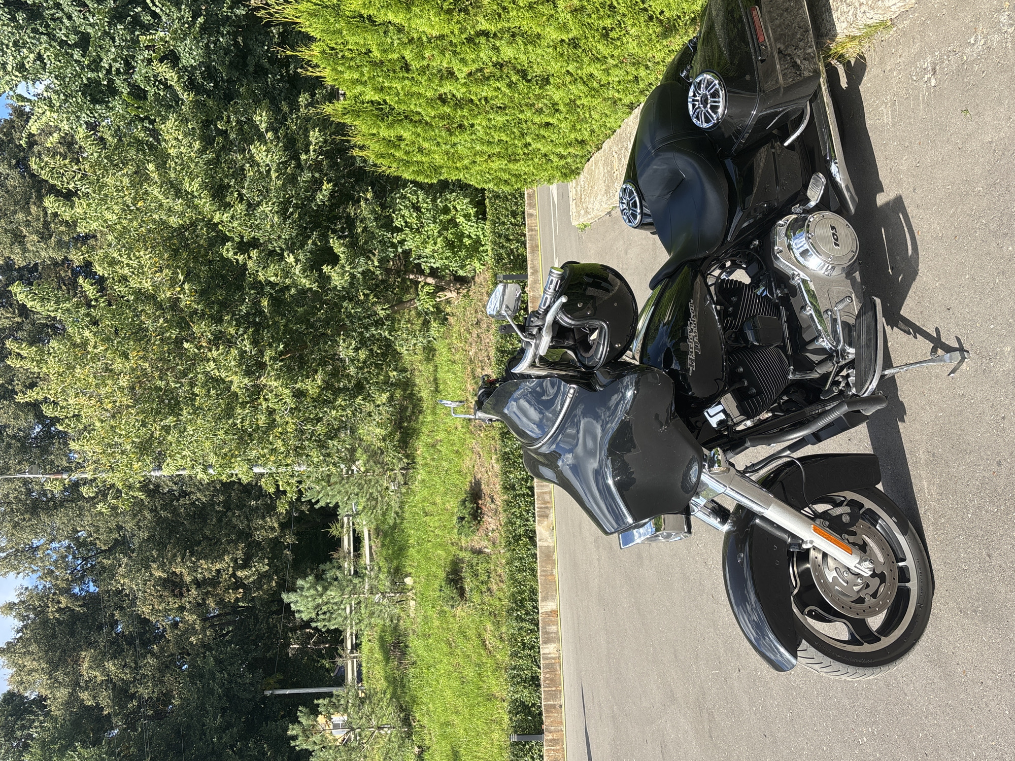 A black Harley-Davidson motorcycle is parked on an asphalt road next to a well-maintained cypress hedge. The motorcycle is facing the viewer's left, with its front wheel slightly turned. The bike has a large black fairing, a black seat, and chrome accents. A black helmet is hanging from the handlebars.
Behind the motorcycle, a grassy slope rises, dotted with small pine trees and larger deciduous trees forming a dense forest in the background. A low stone border separates the road from the grassy area. The scene appears to be a quiet, scenic location, likely a park or a private estate, given the landscaping. The sky is blue with some clouds. The overall impression is one of a peaceful, sunny day. The image does not contain any people. The location is Zvenigorod, Russia.