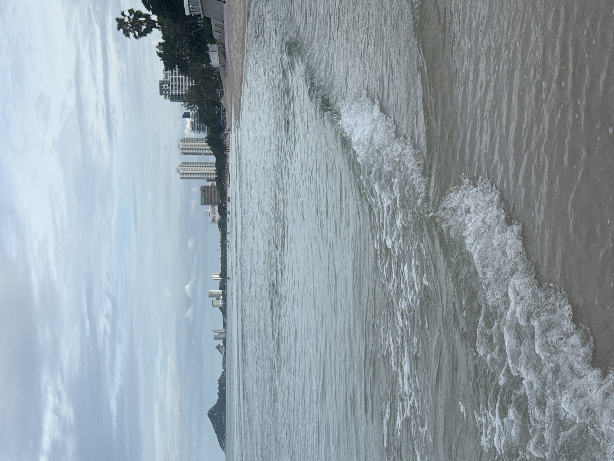 The image shows a view of the beach in Hua Hin City Municipality, Thailand. The foreground is dominated by the ocean, with small waves lapping at the shore. The water is a muted gray-blue, reflecting the overcast sky. In the middle ground, a sandy beach stretches out, with several people visible enjoying the day. Beyond the beach, a line of tall buildings, indicative of a city, rises against the horizon. To the far left, a distinctive mountain formation juts out into the sea. A single palm tree stands prominently on the right side of the image, adding a tropical element to the urban coastal landscape. The overall scene depicts a typical day at a bustling beach town.