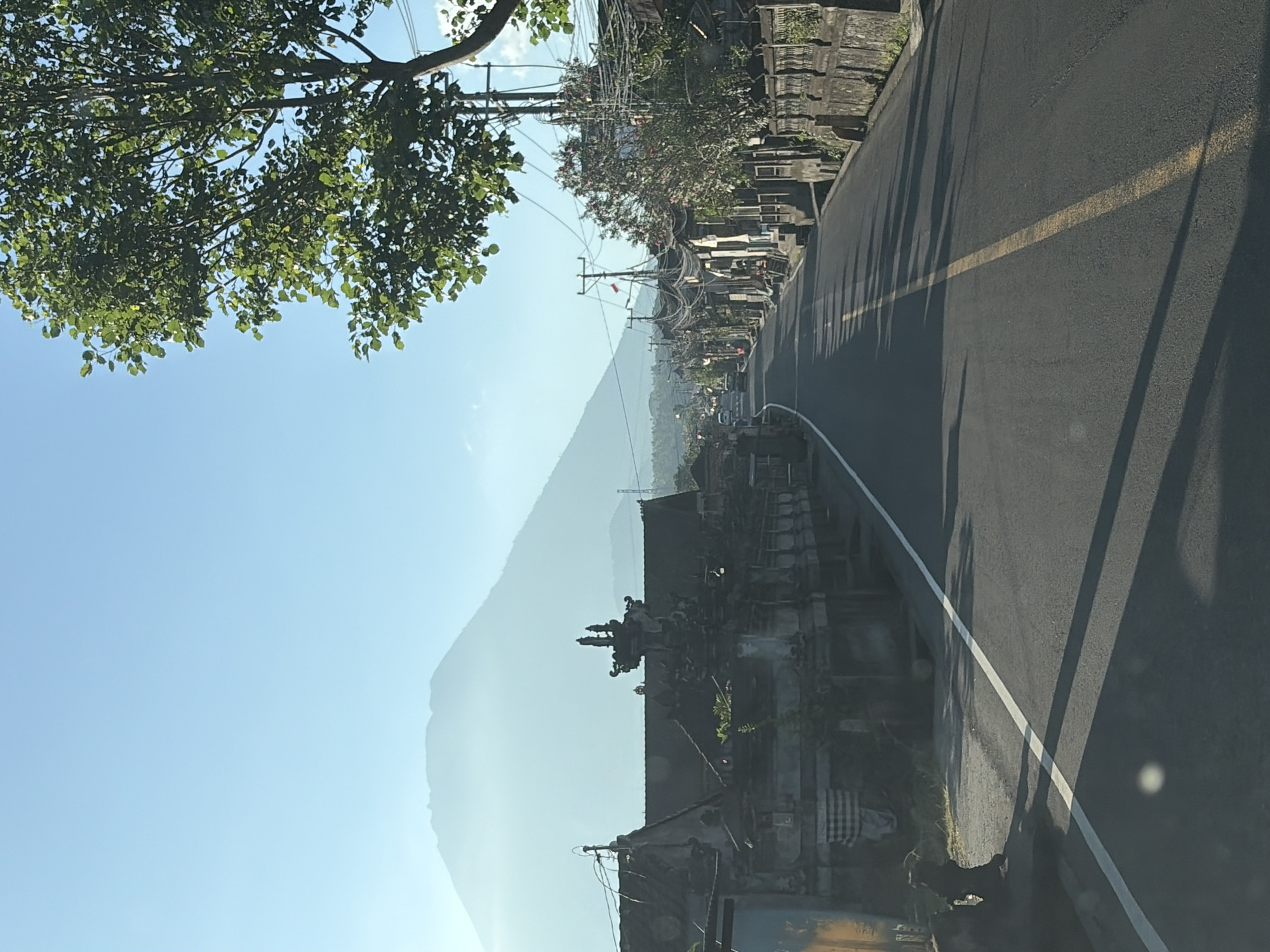 The image captures a road scene in Karangasem, Indonesia, under a clear blue sky. A large, hazy mountain dominates the background, its silhouette softened by the distance. To the left of the road, traditional Balinese architecture is visible, with ornate stone carvings and tiled roofs. A black dog is present on the left side of the road, sitting near a stone wall. The road itself is paved and has a yellow center line. On the right side, a row of buildings with similar architectural styles line the street, interspersed with trees and utility poles carrying numerous wires. The overall impression is a quiet, early morning or late afternoon view of a rural or semi-urban area in Bali.