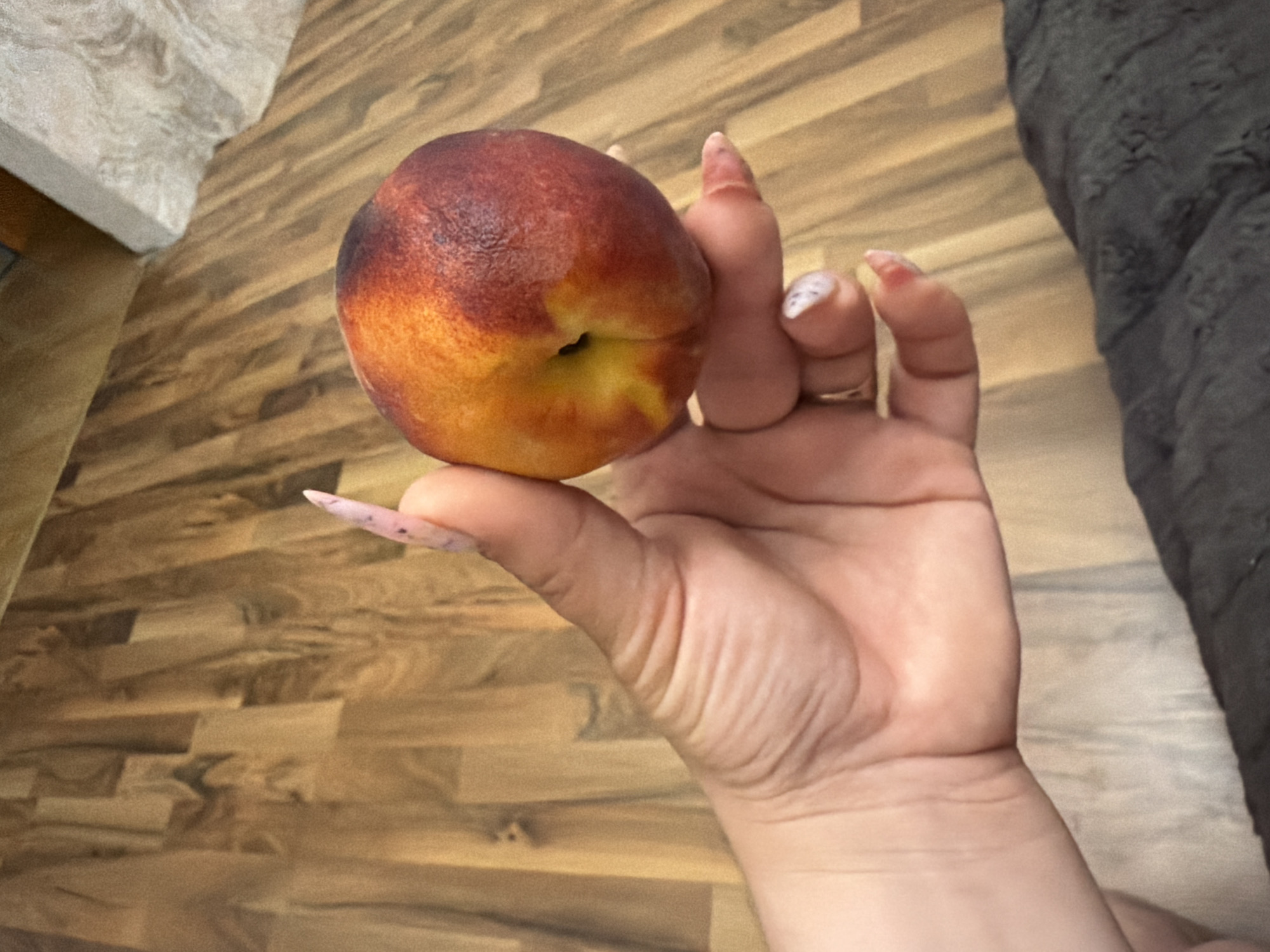A person's hand, with manicured nails, holds a single peach against a backdrop of a wooden floor. The peach is round and shows a gradient of colors, from yellow near the stem to a deep red-orange on the outer portion. The hand is positioned with the palm facing the viewer, fingers curled around the fruit. The wooden floor is composed of light brown planks arranged lengthwise. The lighting is neutral, and the overall scene appears to be indoors. There is no information available to determine the location in the city of Kyiv, Ukraine.