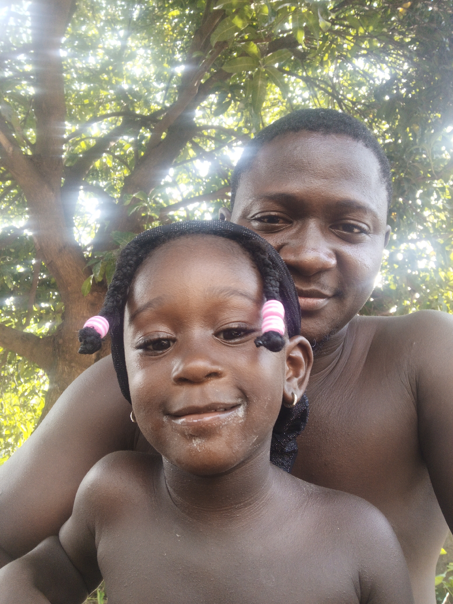 This is a selfie of an adult and a child in a natural, outdoor setting, likely a park or a backyard with a large tree providing shade. The adult, a man with dark skin, is positioned behind the child, with his face slightly out of focus and his bare chest visible. The child, a young girl with dark skin and braided hair adorned with pink and white bands, is in the foreground, looking directly at the camera with a slight smile. Her bare shoulders and upper chest are visible. The background is dominated by the branches and green leaves of a tree, with sunlight filtering through, creating a bright, dappled effect. The overall lighting suggests it is daytime, possibly late morning or afternoon, and the weather appears to be warm and sunny. There are no visible objects or text in the image. The overall mood is casual and intimate, suggesting a moment of shared joy between the two individuals.