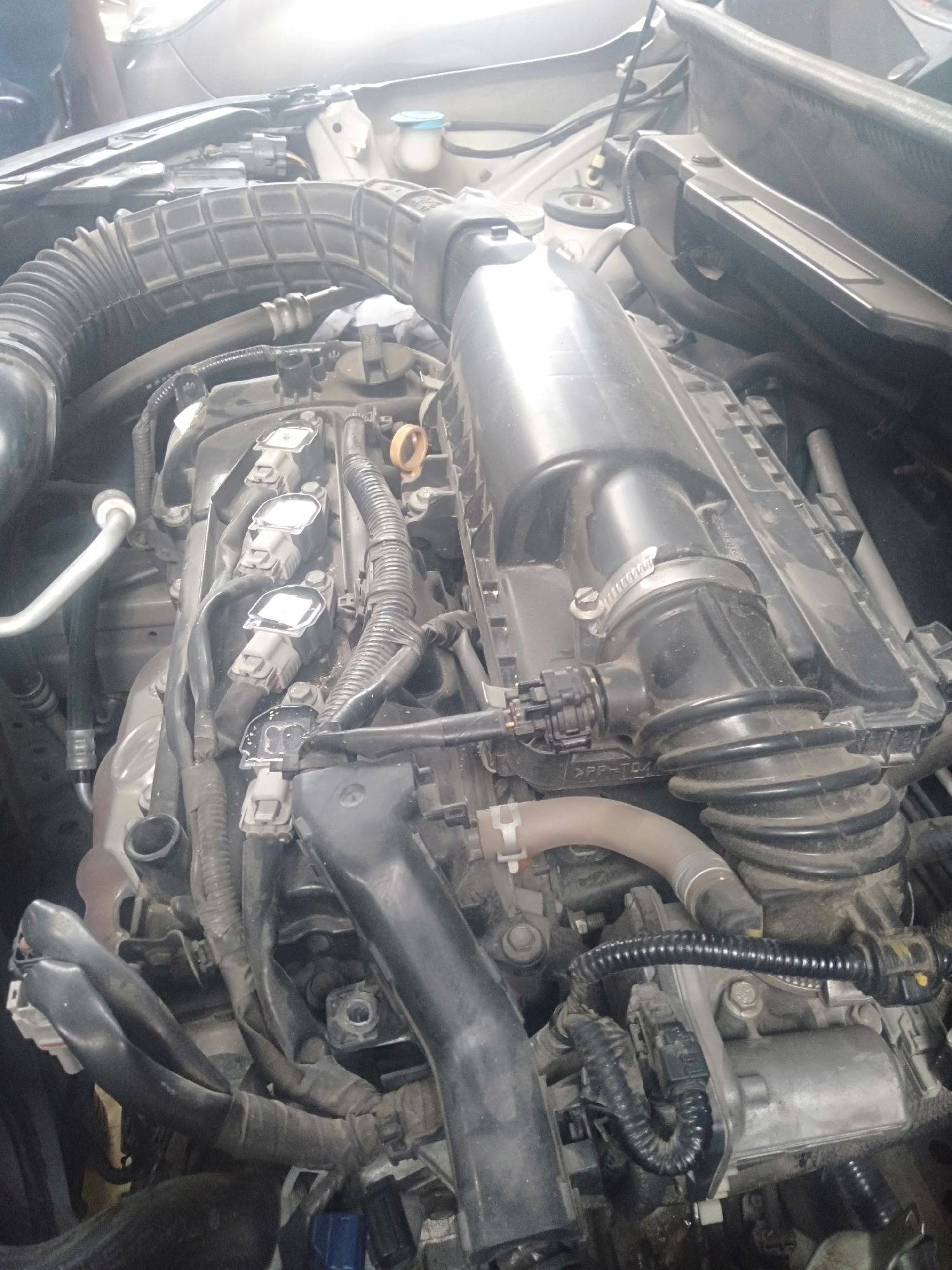 An overhead, medium shot captures the engine bay of a car, which is parked in a residential area in Mojowetan, Indonesia. The engine is comprised of several metallic and plastic components, including the engine block, spark plug wires, and air intake system. The area surrounding the engine is cluttered with wires and hoses. The lighting suggests it is daytime.