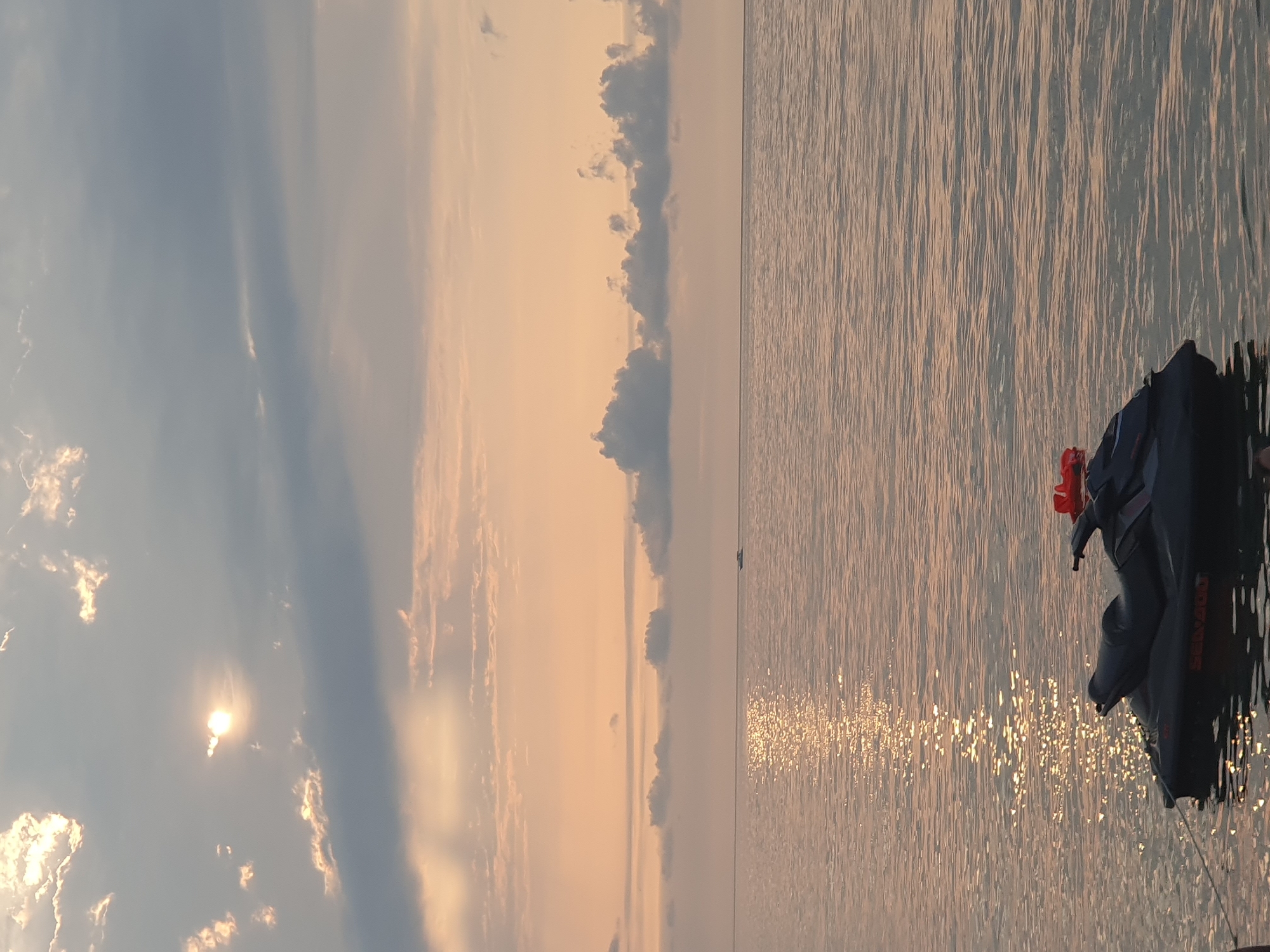 A jet ski is moored in the calm water of the Black Sea, near Batumi, Georgia, during sunset. The sky is filled with a mix of soft orange and grey clouds, with the sun peeking through to cast a shimmering reflection on the water. In the distance, a small boat can be seen on the horizon. The jet ski is dark gray with red accents. The water is smooth, with gentle ripples.