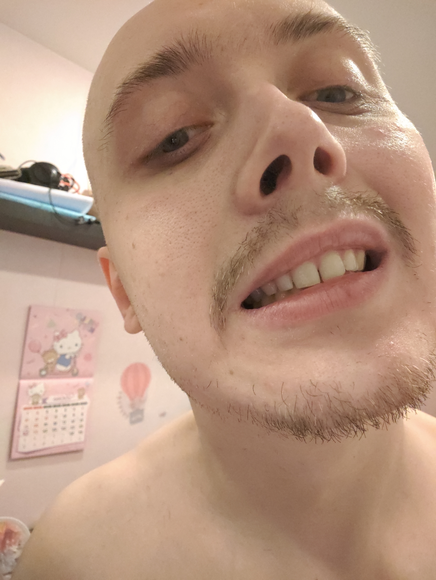 The image is a close-up, low-angle selfie of a person's face, with the camera positioned in front of them. The person is bald and has light stubble on their upper lip and chin. Their eyes are partially closed, and their mouth is slightly open, revealing their teeth. The background is blurred but appears to be a bathroom, with a pink calendar featuring Hello Kitty and a pink hot air balloon decoration visible on the wall. There are also some objects on a shelf in the upper left corner of the image, including headphones. The lighting is warm and appears to be artificial. No specific location in Saint Petersburg, Russia, is identifiable.