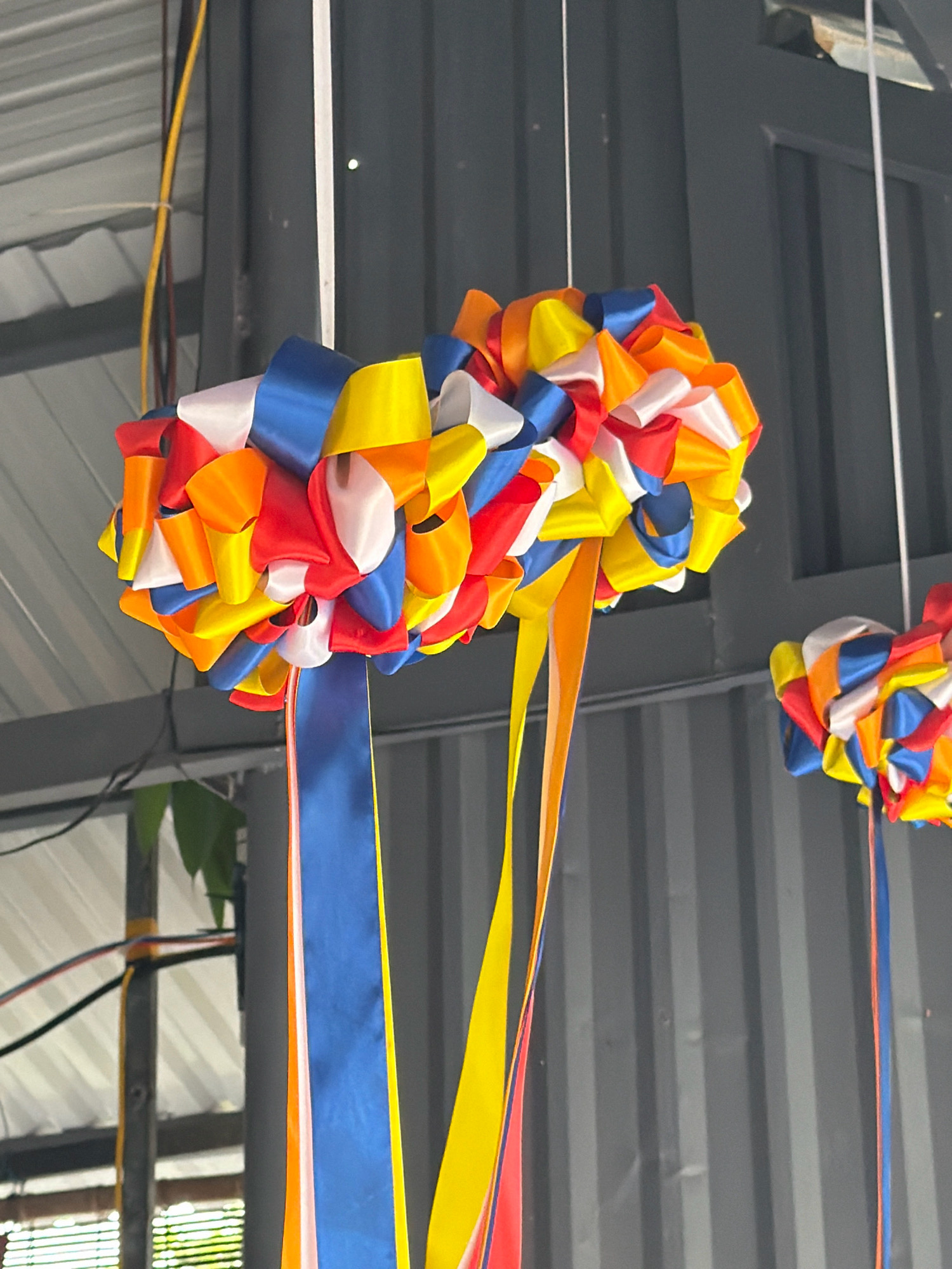Two decorative spheres, made of tightly packed, colorful ribbons in red, orange, yellow, blue, and white, are suspended from the ceiling in what appears to be an indoor market or event space. The spheres are hanging at different heights. The larger sphere, in the foreground, has long ribbons of blue, orange, and yellow hanging down from it. A smaller sphere, partially visible in the background, is also composed of the same vibrant ribbon colors. The background consists of corrugated metal walls and a ribbed ceiling with visible electrical wiring and support structures. The overall environment suggests a utilitarian, possibly temporary, setup. The scene is devoid of any people.