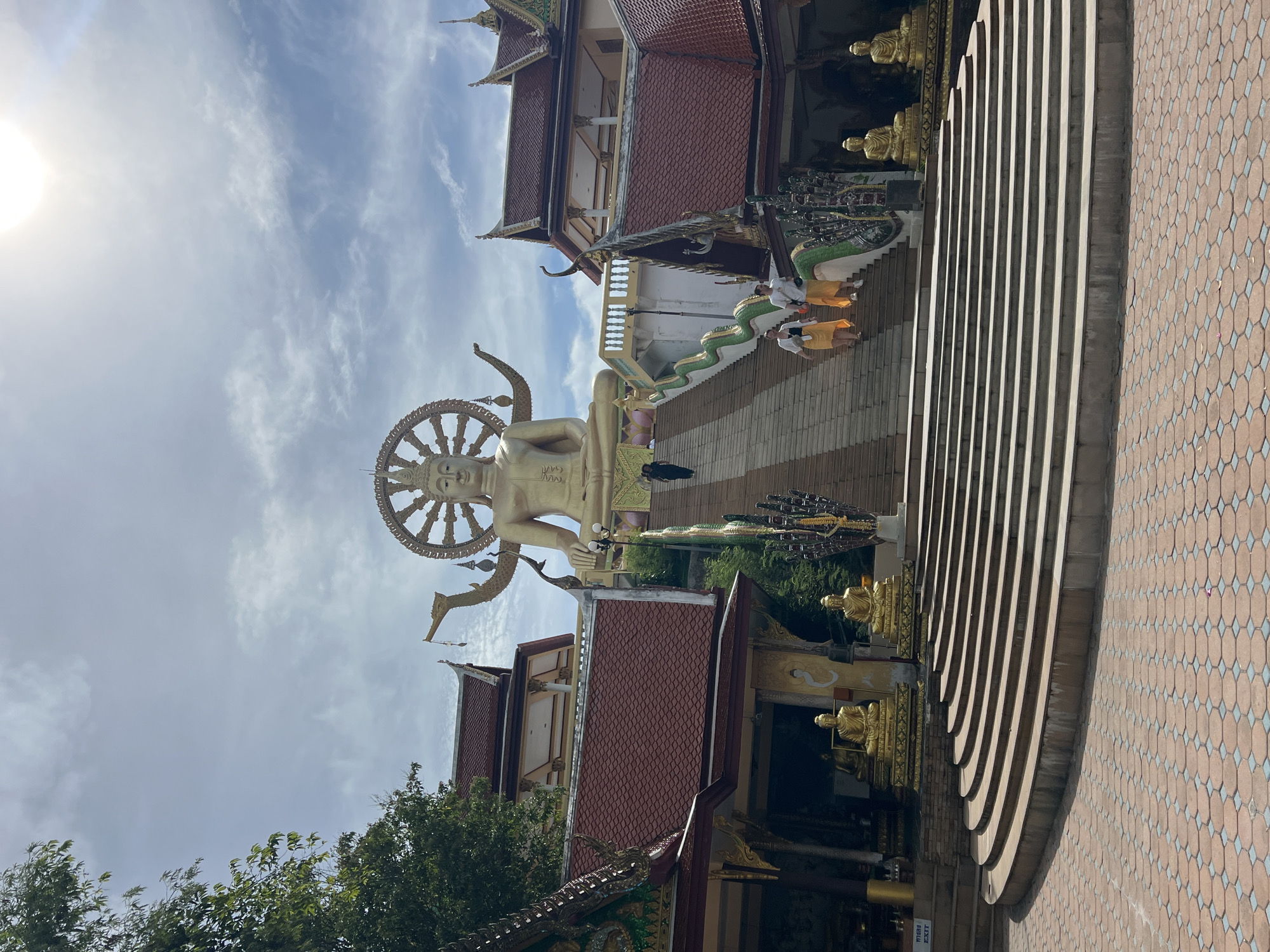 In Baan Plai Laem, Thailand, a large golden Buddha statue sits atop a temple complex under a bright, cloudy sky. The Buddha is depicted with a halo of spokes and multiple arms. In front of the statue, a wide set of stairs ascends towards the temple buildings, which have traditional red tiled roofs and ornate golden accents. Two individuals, dressed in white tops and orange skirts, are walking down the stairs. Another person, dressed in black, walks up the stairs in the background. The lower portion of the image shows a large, semicircular set of stone stairs leading to the main staircase, with a paved courtyard in the foreground. Smaller golden Buddha statues are placed at intervals around the temple grounds.