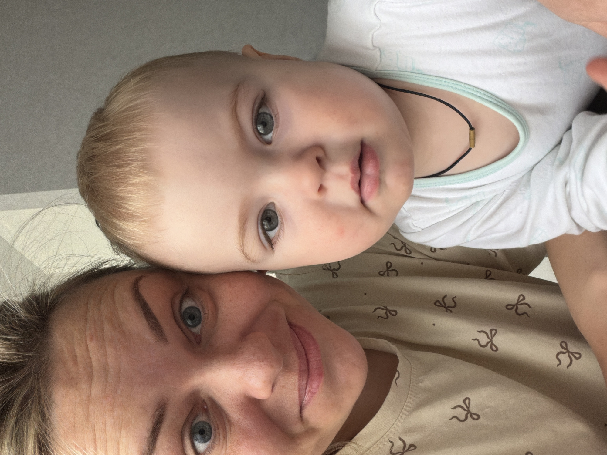 This close-up, vertically oriented photograph captures an intimate moment between an adult woman and a baby or toddler, likely indoors in Votkinsk, Russia. The image is taken from a high angle, looking down at both subjects.
In the lower portion of the frame, the woman, presumably the mother, has light brown hair and blue eyes that look directly at the camera. She displays a slight, closed-mouth smile, and noticeable horizontal lines are visible on her forehead. She is wearing a light beige or brown top patterned with numerous small, dark brown bow shapes.
Above her, resting their head on the woman's, is a baby or toddler. The child has very fair, light blonde hair and large, striking greyish-blue eyes that also engage directly with the viewer. The child's cheeks are rosy, and their expression is calm and neutral, perhaps a little curious. They are dressed in a white long-sleeved top with a light blue trim around the neck, and wear a thin black cord necklace adorned with a small, gold-colored cylindrical bead.
The background is a plain, light-colored surface, possibly a wall or ceiling, indicating an indoor setting. The overall atmosphere of the photograph is tender and peaceful, highlighting the close bond between the two individuals. No visible text is present in the image.