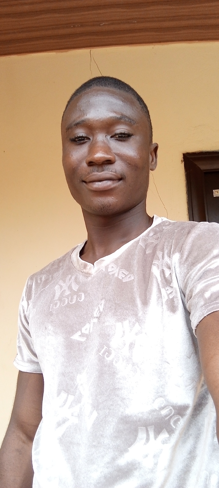 A young adult male of African descent is captured in a head-and-torso shot, facing the camera with a slight smile. He has short, dark hair, minimal facial hair, and visible scars on his right cheek. He wears a light beige or off-white short-sleeved t-shirt featuring embossed or textured patterns, including the word "GUCCI" and a logo resembling the New York Yankees "NY" emblem. The subject is positioned against a plain, light tan-colored wall. Above his head, a dark brown wooden beam runs horizontally across the top of the frame. A thin, jagged crack is visible descending from the beam into the wall directly above the subject. To the far right, a dark brown door frame is partially visible, containing a dark panel with a small, white rectangular sticker or label affixed to it. The scene takes place within a built environment in the city of Dzodze, Ghana.