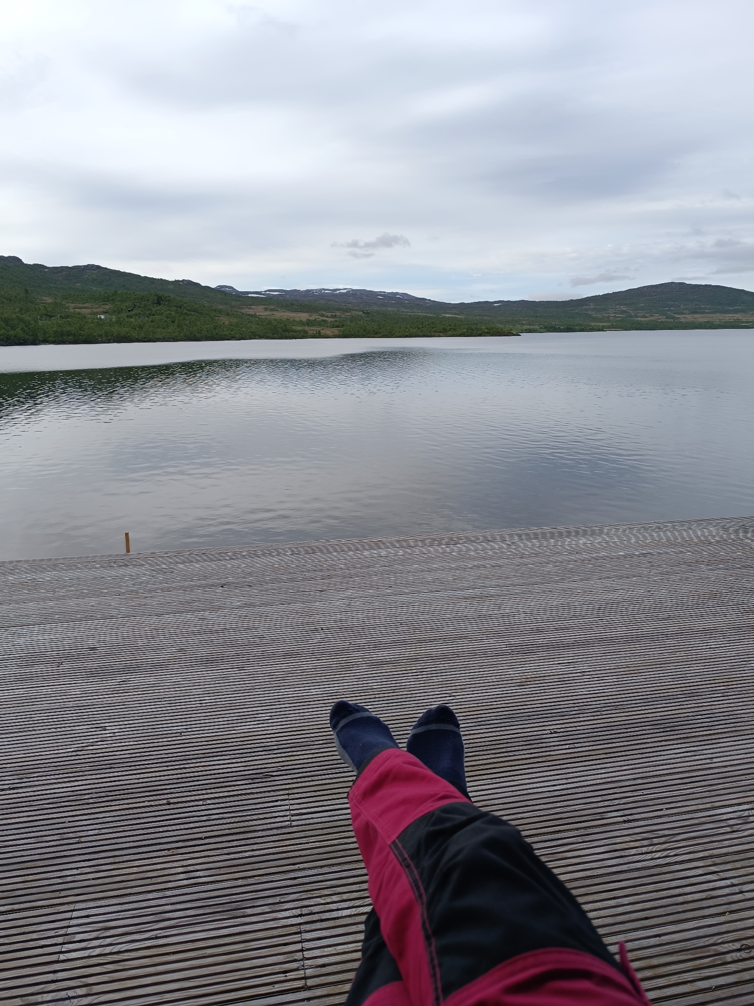 The image shows a person's legs and feet resting on a wooden deck. The person is wearing dark blue socks and red and black pants. The wooden deck is made of many narrow planks laid horizontally across the frame. In the background, a large body of water is visible, with calm ripples on its surface. Beyond the water, there are rolling green hills covered with trees. The sky is overcast with pale grey clouds. The location is identified as Hovden, Norway.