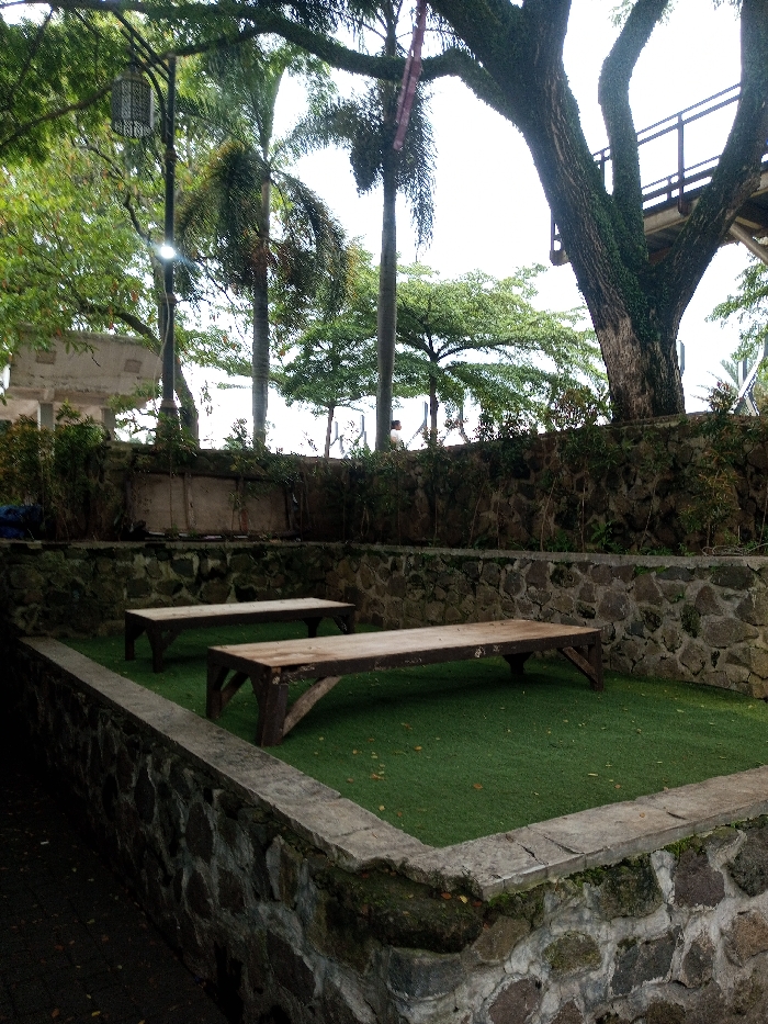Two wooden benches sit on a patch of artificial green turf within a low stone-walled enclosure. The wall is constructed from irregularly shaped stones. Behind the enclosure, another higher wall is visible. Trees, including palms and a large tree with a thick, mossy trunk, provide a backdrop. Above, a bridge structure can be seen through the branches. A lamppost stands to the left, adding a touch of artificial light to the scene.
