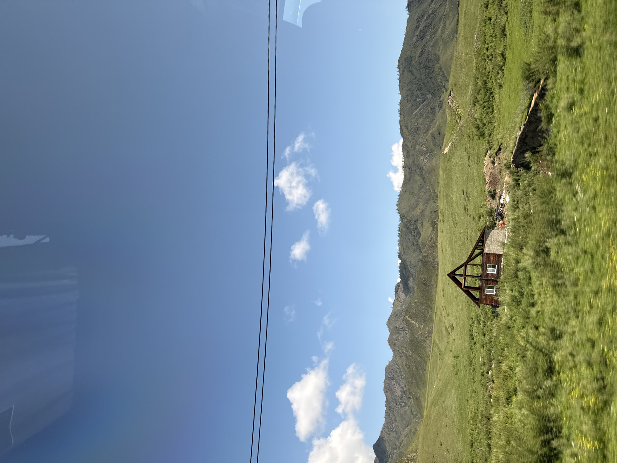 A clear, bright day is captured in this image from Tuekta, Russia. The sky is a vibrant blue with a few scattered white clouds. In the foreground, a green, grassy field stretches out, with a partially constructed wooden building located in the middle. The building appears to be a house with visible windows and a wooden frame, suggesting it is either under construction or in disrepair. Behind the field, rolling green hills and mountains rise, their peaks partially obscured by the distance. Power lines are visible in the upper portion of the image, stretching horizontally across the frame. The overall impression is one of a rural, undeveloped landscape under a sunny sky.