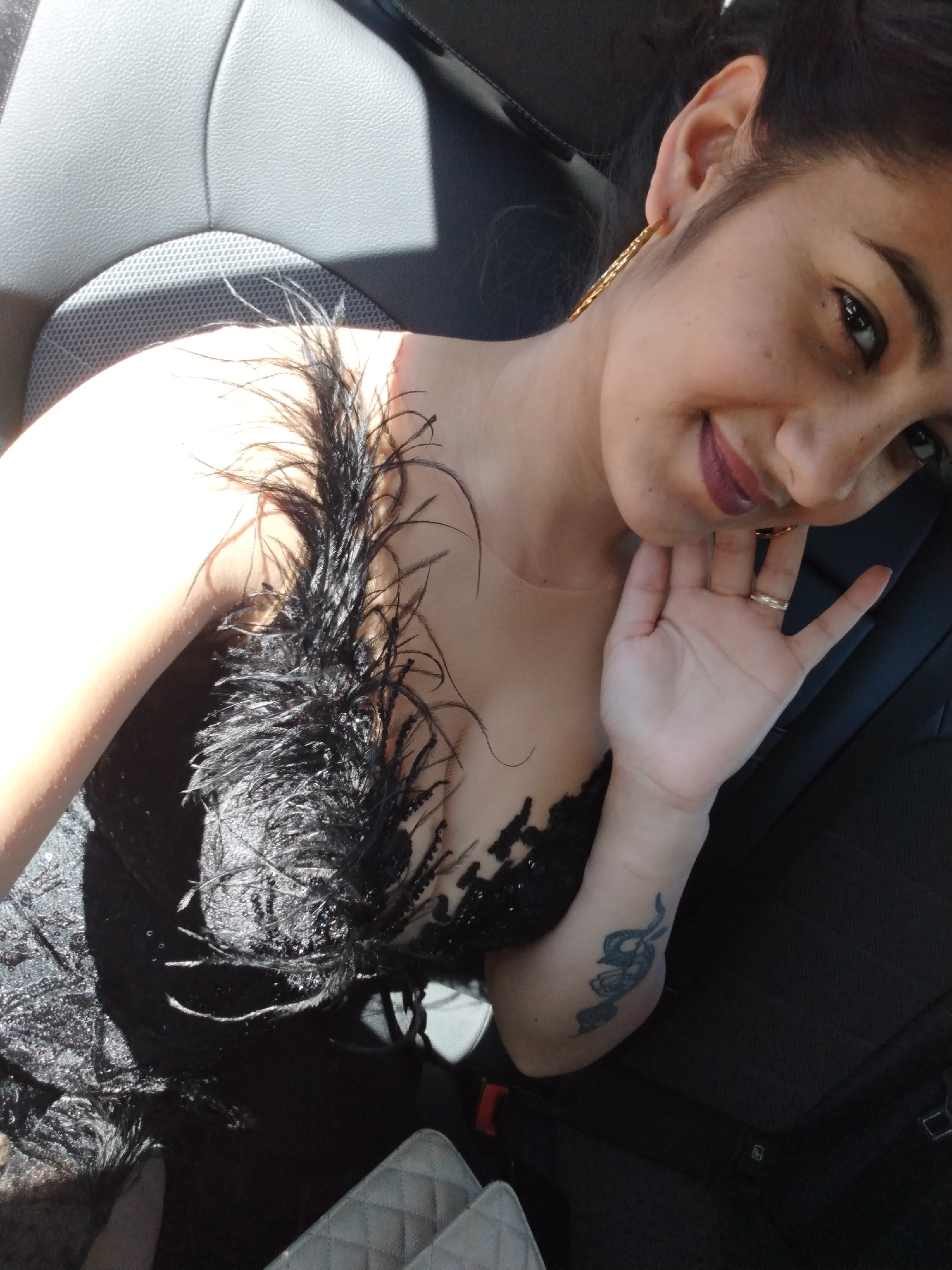 A woman with dark hair is seen from above, seated in the backseat of a car. She is wearing a black, strapless dress adorned with black feathers. A tattoo is visible on her left forearm. The car's interior is dark, with a gray seat visible behind her. The lighting suggests it is daytime, possibly with sunlight filtering into the car.