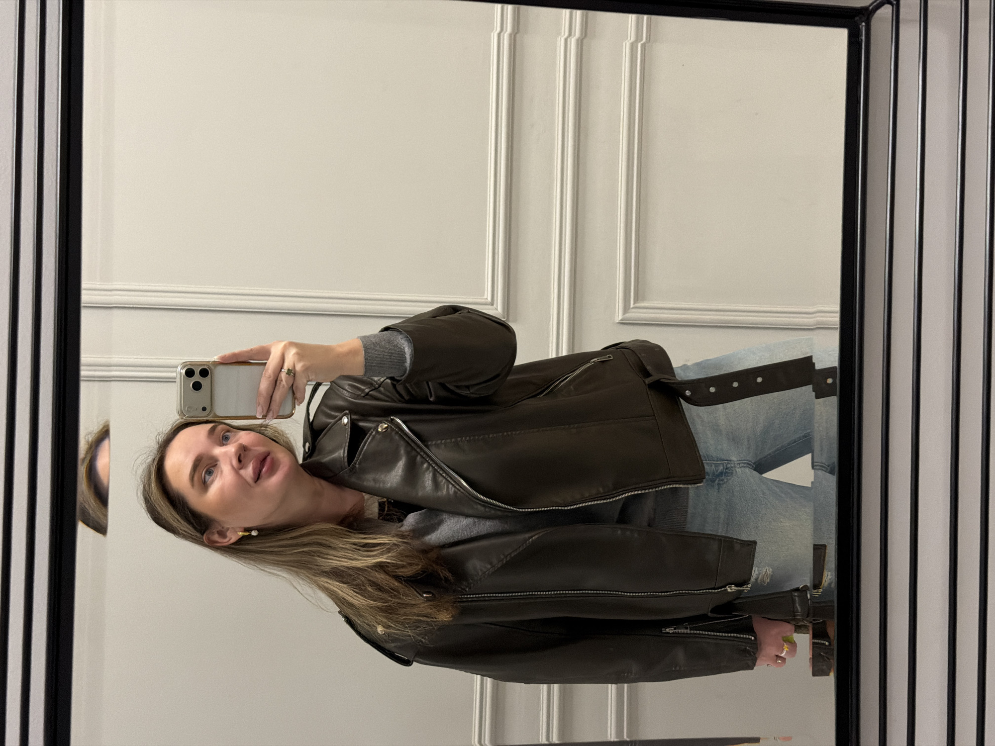 The image is a mirror selfie of a Caucasian woman with blonde hair and blue eyes. She is holding a smartphone in her right hand, taking a picture of herself in the mirror. She is wearing a grey sweater, light blue jeans, and a dark brown leather jacket. The background is a light grey wall with vertical decorative panels. The mirror has a dark frame with additional vertical lines on the outer edges. The location is difficult to determine from the image. Based solely on the visual information, there is no definitive way to identify this location as Moscow, Russia.