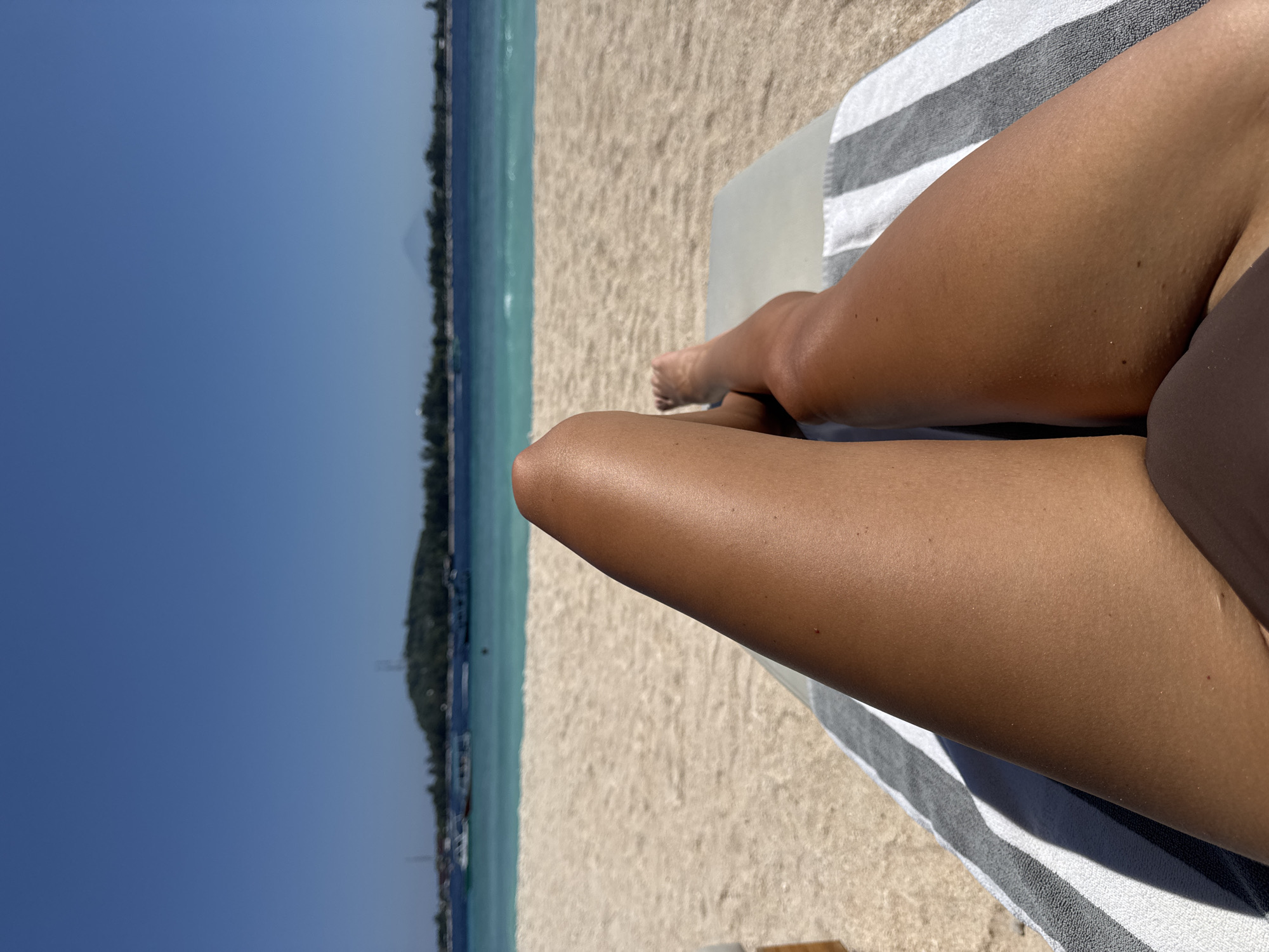 The image presents a view of a person reclining on a beach, likely on a lounge chair, in the location of Unknown, Indonesia. The person's tanned legs are prominently featured, resting on a striped towel. The background includes a sandy beach and turquoise water, with a coastline featuring trees and possibly buildings in the distance. The sky is clear and blue.