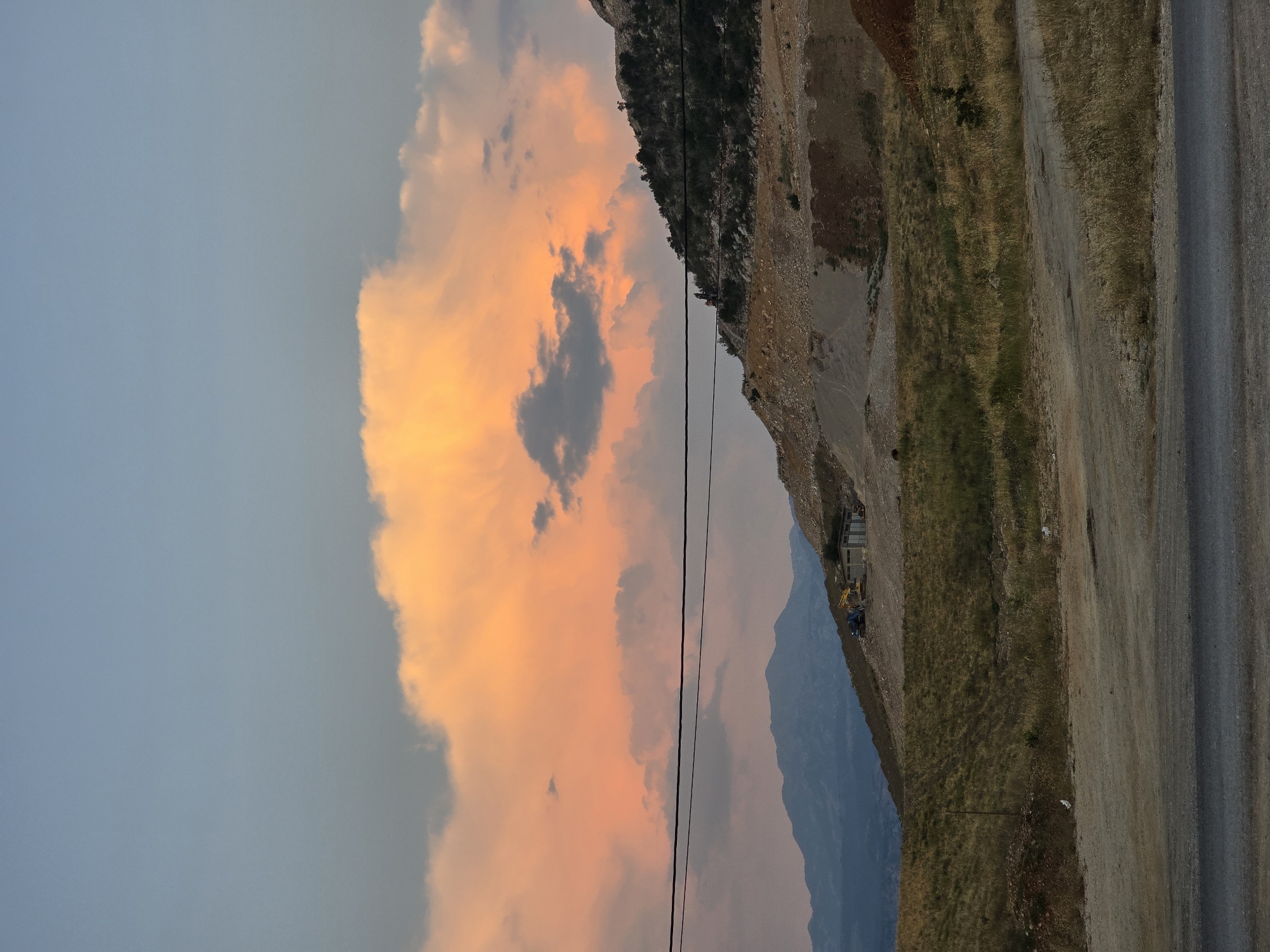 In Orljevo, Montenegro, at dusk, the sky is a gradient of pale blue and orange. A large, illuminated cloud dominates the upper center of the frame, tinged with warm hues of pink and yellow. Below the sky, a power line stretches horizontally across the scene. A rugged, mountainous terrain occupies the middle ground. The slopes are sparsely vegetated with dry grass and scattered rocks, indicative of arid conditions. Construction is underway on one of the hills, with several pieces of heavy machinery, including yellow excavators and a blue digger, present. A small building with a gray roof is also visible at the construction site. In the background, distant, hazy blue mountains form a layered backdrop. The foreground features a dirt road or path, transitioning into an asphalt road at the bottom of the frame. The overall impression is one of a developing landscape under a dramatic sky.