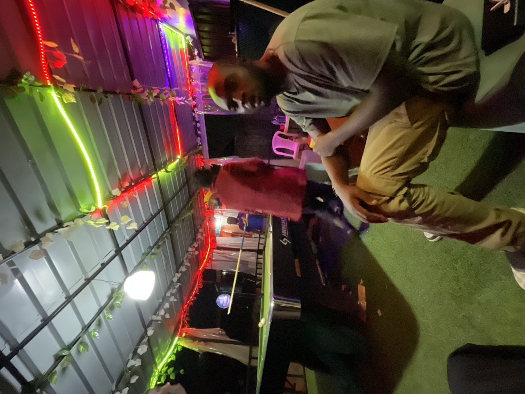 The image shows a nighttime scene in Barakin Akawo, Nigeria. A man in a green t-shirt and tan pants is bending over, partially obscuring the view. Another person in a red shirt stands further back. A pool table and what appears to be a sound system speaker are visible. The background is decorated with strings of green, red, and purple LED lights, and the ceiling is made of corrugated metal.