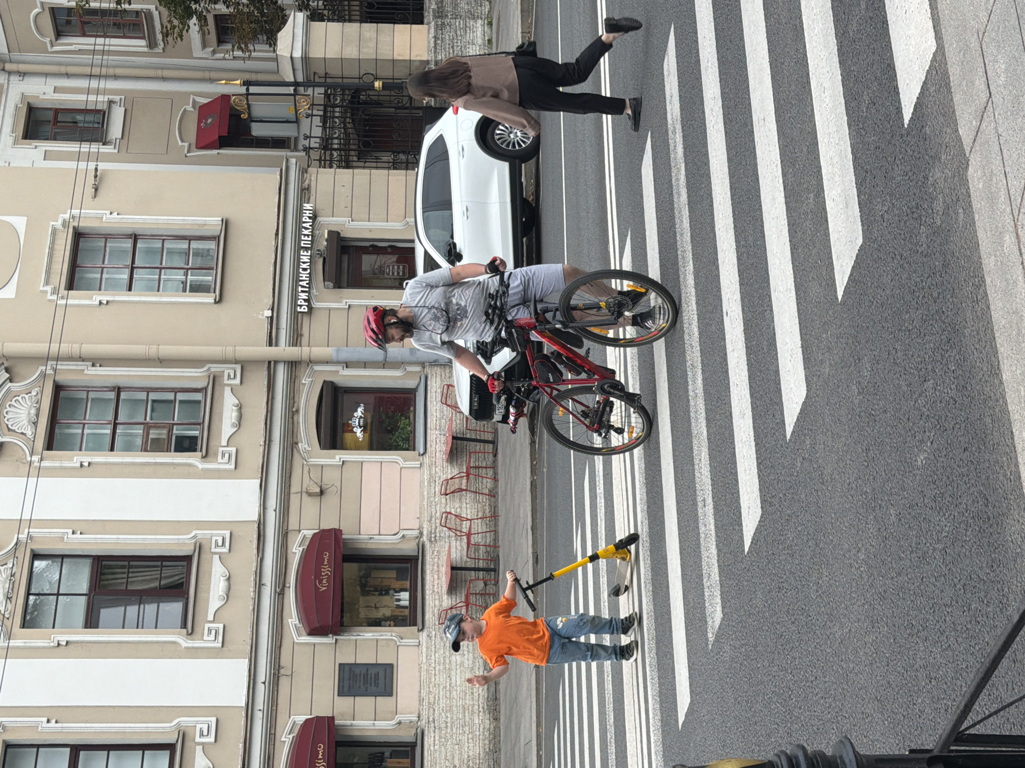 On a cloudy day in Saint Petersburg, Russia, a young boy wearing an orange t-shirt and jeans is depicted standing on a crosswalk with a yellow and black scooter. He is waving with his right hand. To his right, a man wearing a red helmet, grey t-shirt, and light grey shorts is on a red bicycle, facing away from the camera. Beside him and slightly behind, a woman with dark hair, wearing a brown jacket and black pants, is walking to the right. A white SUV is parked in the background, and the facade of a building with multiple windows and awnings is visible. The building has a sign with Cyrillic text. The street is paved with asphalt and marked with white pedestrian crossing lines.