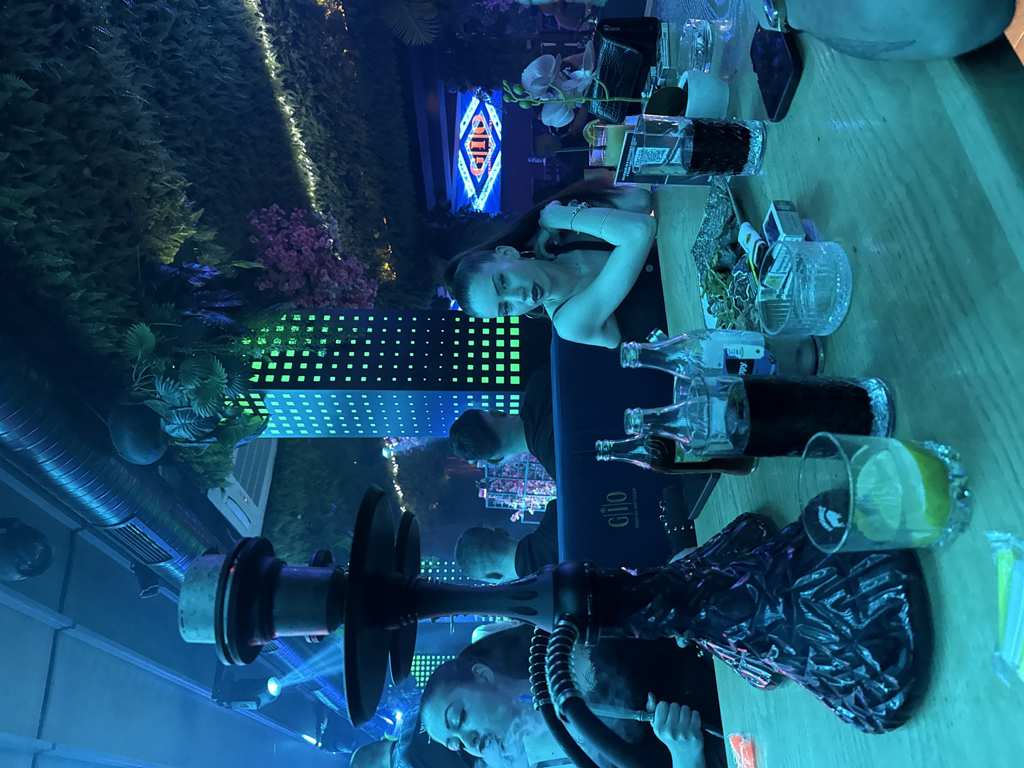 The image captures a dimly lit indoor scene, likely a bar or lounge, characterized by vibrant blue and green artificial lighting. The original photograph is rotated 90 degrees clockwise.
In the mid-ground on the right, a woman with dark hair pulled back is leaning casually on a light-colored wooden bar or table, looking directly towards the camera. She is wearing a dark top and a bracelet on her left wrist. In the foreground, on the left, a large, dark hookah is prominent, with a person (partially visible) exhaling a cloud of smoke from it.
The bar surface is cluttered with various objects, including several glass bottles, one of which has a label clearly reading "CLIO PREMIA BESTSELLER". A glass containing a drink with a lemon or lime slice is also visible, along with a small white orchid plant in a pot, a black wallet, and an ashtray-like glass dish holding matches and other small items.
The background features lush artificial greenery, resembling ferns and other plants, covering a wall. Clusters of pink flowers and panels with illuminated green square patterns are integrated into the decor. A neon sign in the distant background spells out "CLIO". Further back, at least two men are visible, one appearing to be behind the bar. Exposed metallic air ducts are visible on the ceiling.
The scene suggests a lively, social nighttime setting where patrons are enjoying drinks and hookah in a stylish, mood-lit environment.