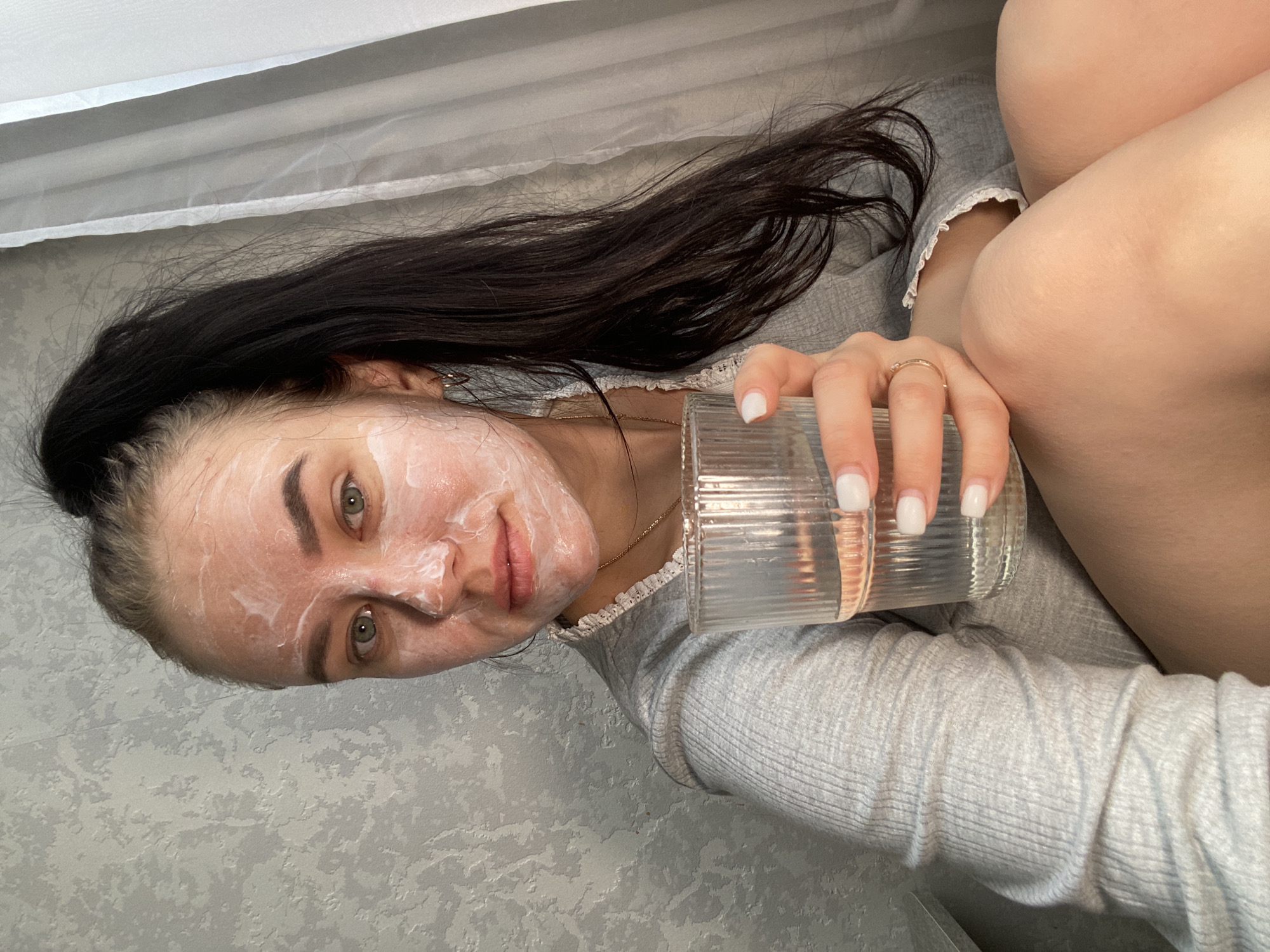 A woman with a facial mask is holding a glass of water. She has dark hair pulled back and is wearing a gray long-sleeved shirt. The background includes a gray wall and a white draped object. There is no evidence in the image to support the location being in Chaikovsky, Russia.