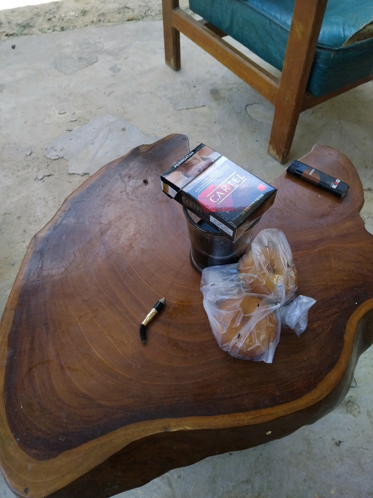 A wooden coffee table in Andoolo, Indonesia, displays several objects: a pack of "Cartel" kretek filter cigarettes, a metal ashtray, a lighter, two pieces of bread in a plastic bag, and a single cigarette butt. A portion of a wooden chair with blue upholstery is visible in the background, set against a concrete floor.