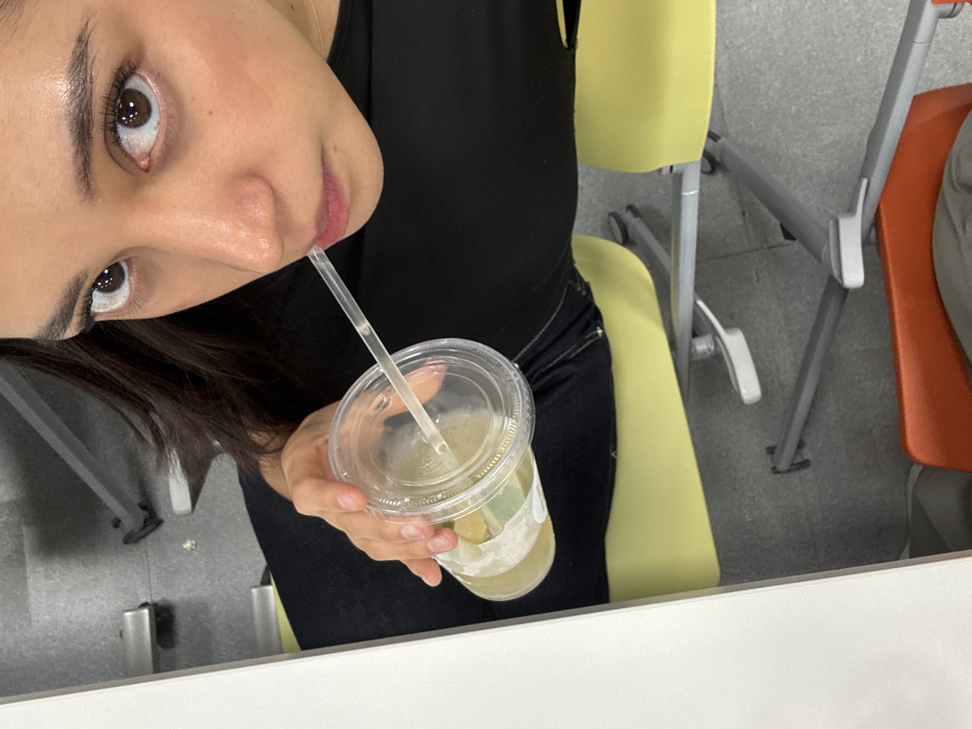 A woman is holding a plastic cup with a drink and a straw. The drink appears to be light-colored with some solids in it, possibly fruit. She is drinking from the straw. She has dark hair and dark eyes. She is wearing a black top and dark pants.
The background includes chairs with yellow and orange seats. The floor appears to be carpeted with a grey color. The scene appears to be indoors, possibly a school or office in Puebla, Mexico.