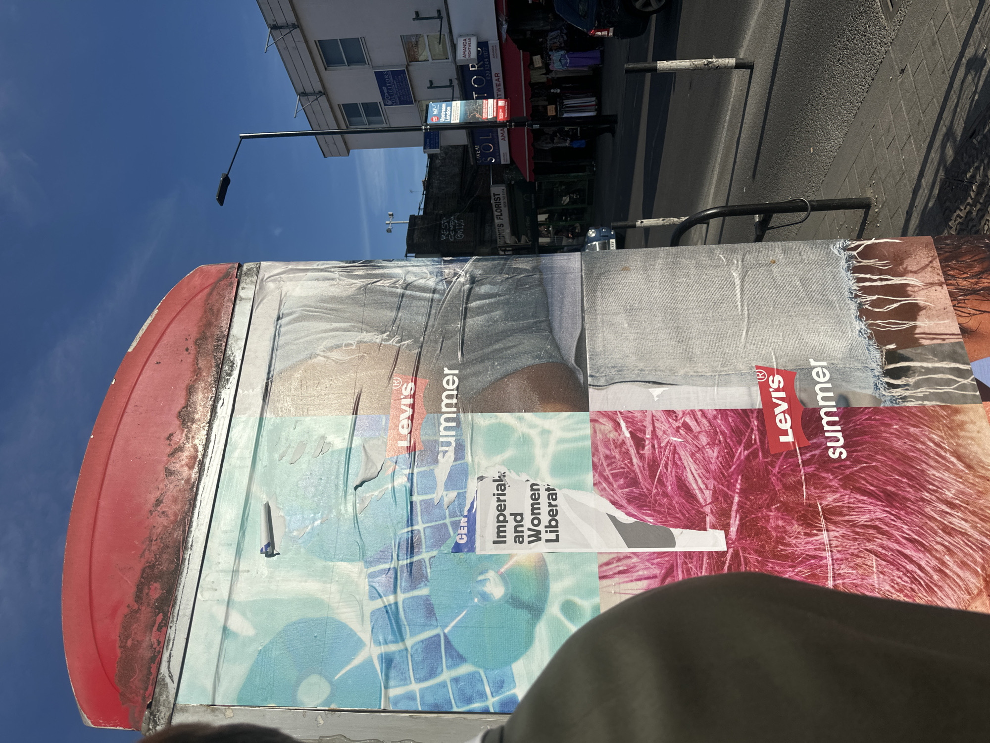 This image shows a bulletin board covered with posters, including Levi's Summer advertising. Behind it is a street in London, United Kingdom. Several storefronts are visible, including "INT'S FLORIST," "Upstream London," "Caveat Solicitors," "Amanda Nightwear," and "Topswear." A streetlight is also visible. The sky is blue.