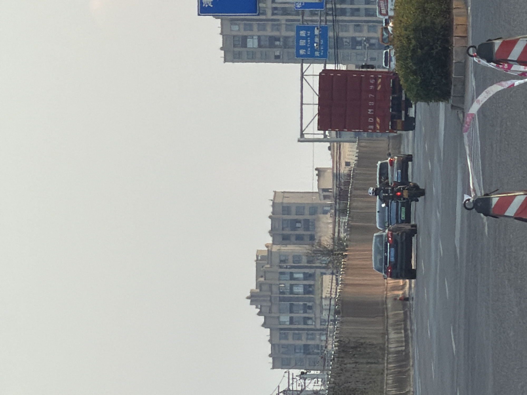 An eye-level, medium shot captures a street scene in Jiangjin, China, under an overcast sky. The focus is on a road with multiple lanes, bordered by a concrete barrier on the left and a traffic island with dense foliage on the right. In the foreground, traffic cones with red and white reflective tape are partially visible. Several vehicles are in motion: a motorcycle rider in black, wearing a helmet and black jacket, is moving away from the camera. A dark-colored sedan follows closely behind the motorcycle, and further ahead, a large red truck with white lettering, possibly indicating a license plate or identification, is also moving away. Beyond the road, buildings of varying heights and architectural styles are present, including a large, multi-story structure with a distinctive stepped facade. Traffic signs, some with Chinese characters, are visible on poles. The overall atmosphere is that of a typical urban environment during daylight hours.