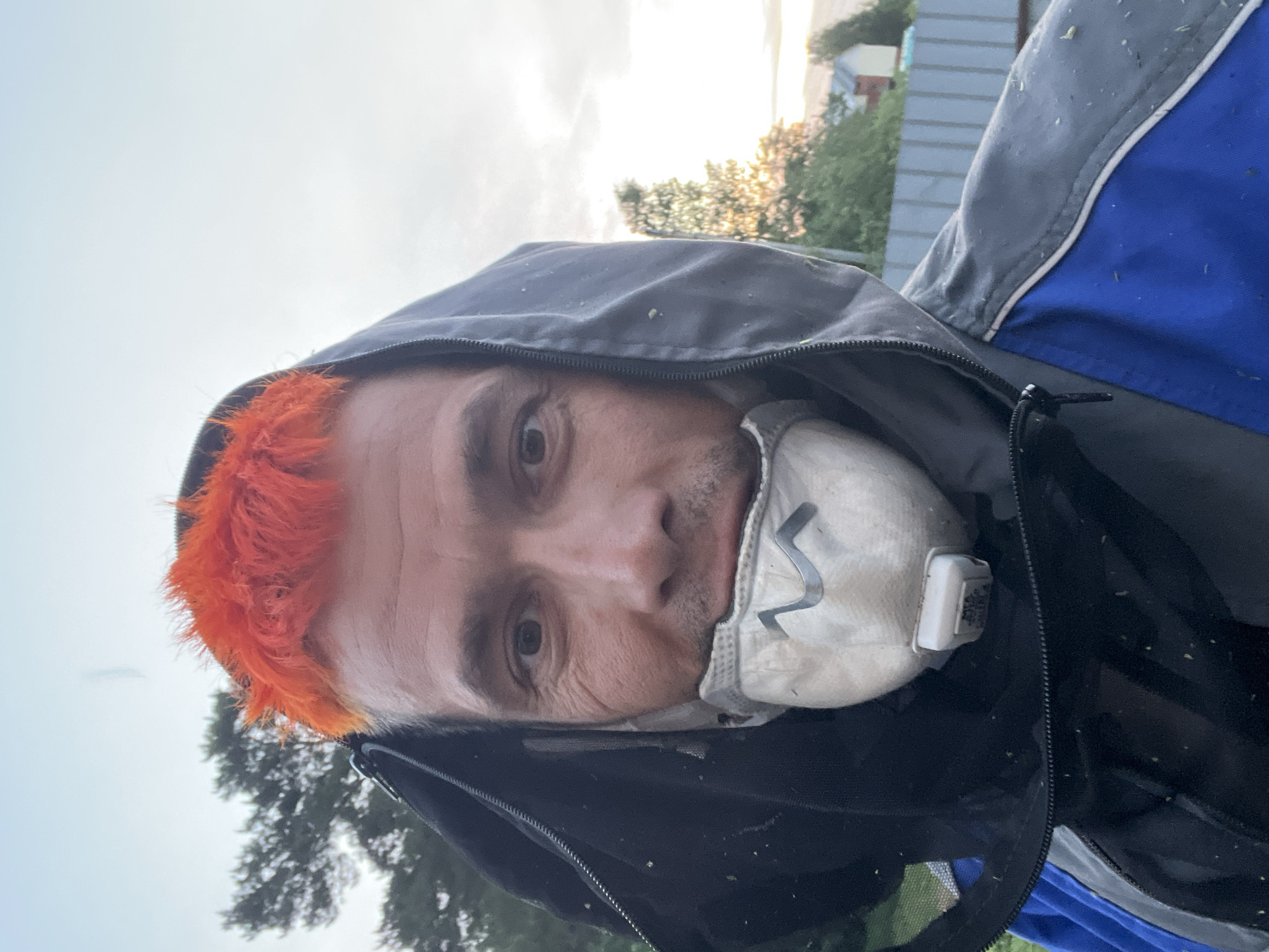 A close-up, slightly angled, medium shot shows a person with orange hair wearing a dark-colored hooded jacket and a white respirator mask. The person's face is visible, showing their eyes, nose, and mouth obscured by the mask. The hood is up, partially covering the hair. The jacket has blue and grey sections. In the background, there are trees, a fence, and a building under an overcast sky. The location may be the city of Chaikovsky, Russia, but there is no visual confirmation of the location, which makes it impossible to confirm with only this image.