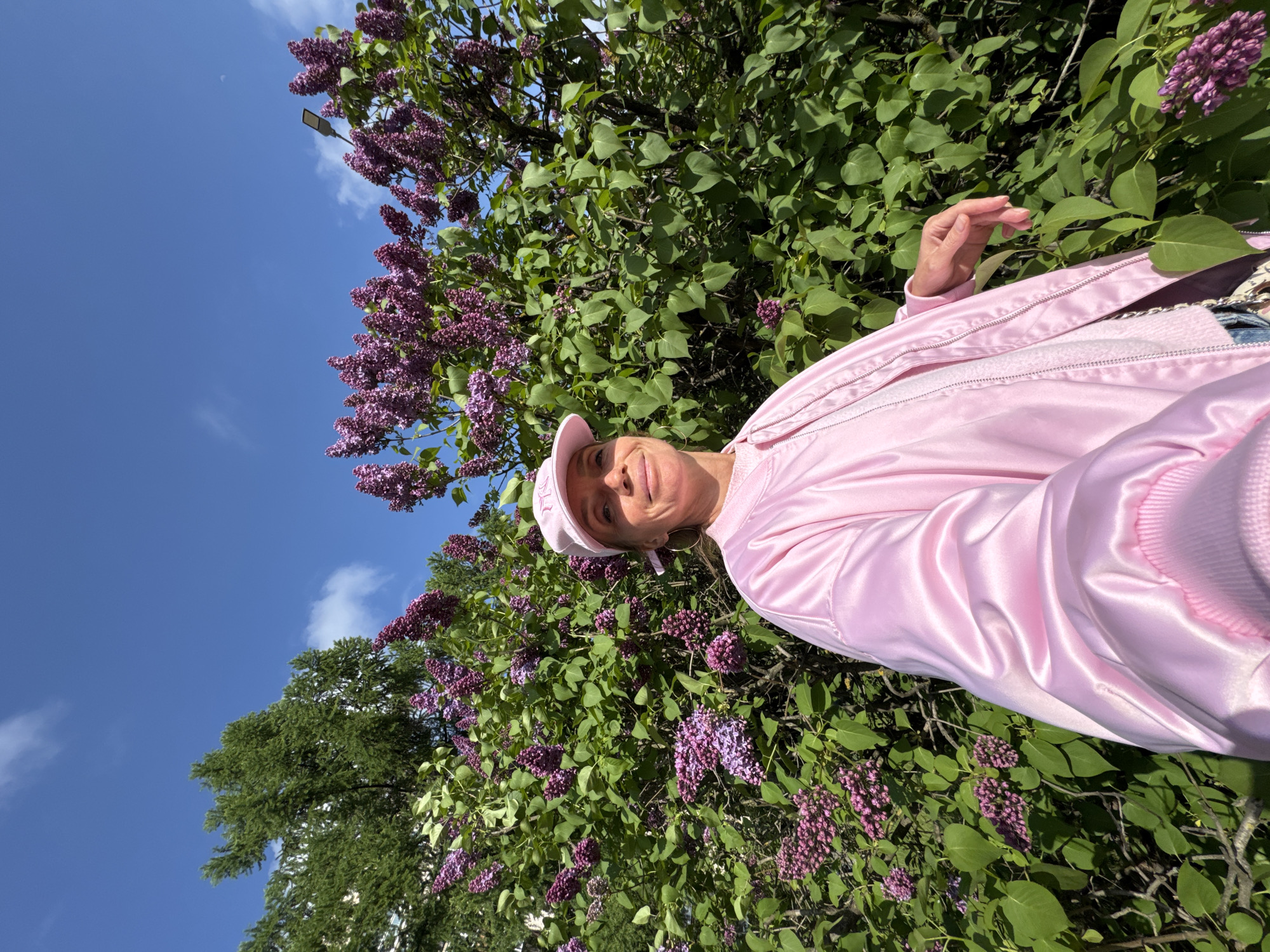 A woman wearing a pink jacket and cap stands in front of a lilac bush with purple flowers, with a clear blue sky and a few clouds visible in the background. The location is likely Saint Petersburg, Russia, given the prevalence of lilac bushes in the city during spring.