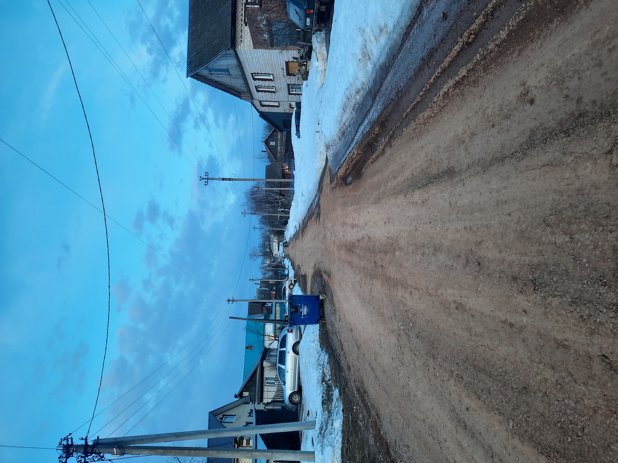The image depicts a street scene in Krasnogorskoye, Russia. The street is unpaved and appears muddy, with melting snow present along the sides. Buildings line the street, including residential structures with brick and wooden facades. Utility poles with power lines run along the side of the street, against a blue sky with some cloud cover. A light-colored car is parked near a blue trash container.
