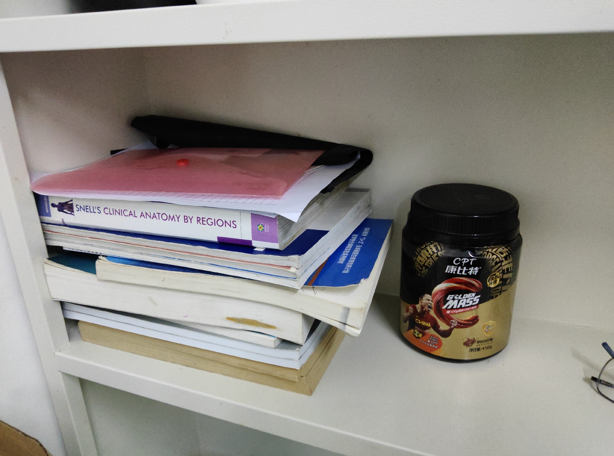 The image shows a shelf in what appears to be a home or office environment. A stack of books, with the title "SNELL'S CLINICAL ANATOMY BY REGIONS" visible on one of the top books, is on the left side of the shelf. Next to the books is a black container with a gold label, featuring text in English and Chinese. The label includes the brand name "CPT" and "GOLDEN MASS" and an image of a person wearing a red sports jersey with "CHINA" written on it. The container is a dietary supplement product. On the far right of the shelf, a pair of glasses is visible. The background is a plain, light-colored wall. The location is not specified in the image.