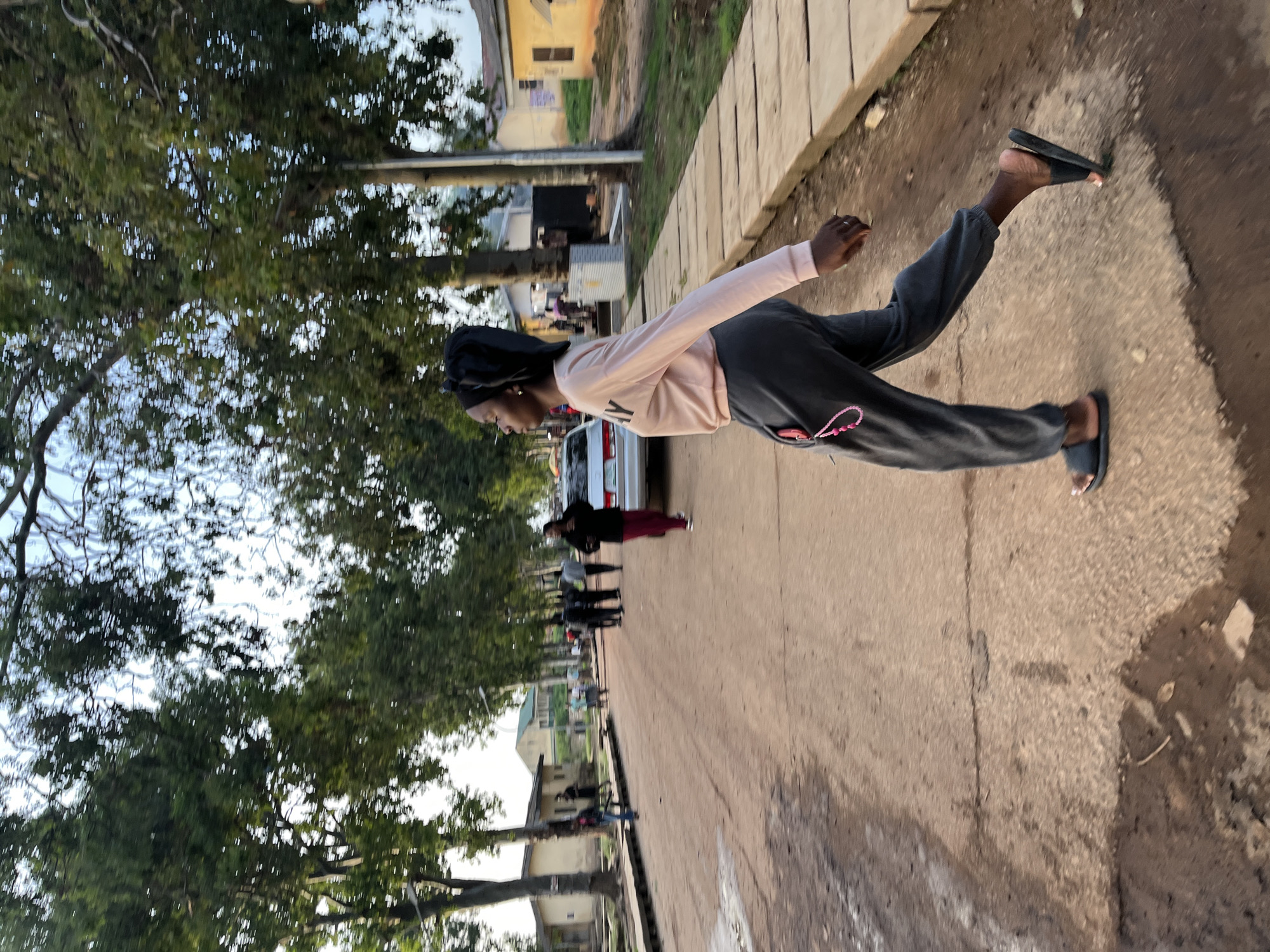This image, set in Rafin Par, Nigeria, depicts a street scene with a young Black woman prominently featured in the foreground, captured mid-stride. She is walking from left to right across the frame, wearing a light pink long-sleeved top that has a partial dark letter (possibly 'Y' or 'V') visible on the chest, dark grey track pants with a pink string or keychain hanging from a pocket, and black slip-on sandals. Her hair is covered by a dark headscarf.
The street itself appears to be unpaved, with a paved sidewalk or curb running along the right side. The background features several other individuals scattered along the street, some standing and others seemingly walking. A white car is parked in the mid-ground, near a person wearing a black top and red pants. Residential buildings with light-colored walls are visible on either side of the road, and the street is lined with numerous large green trees, suggesting a verdant environment.
The lighting indicates late afternoon or early evening, with soft, ambient light illuminating the scene and creating subtle shadows. The overall atmosphere is one of everyday activity on a typical street. No distinct events or strong emotions are evident; the subjects appear to be going about their daily routines.