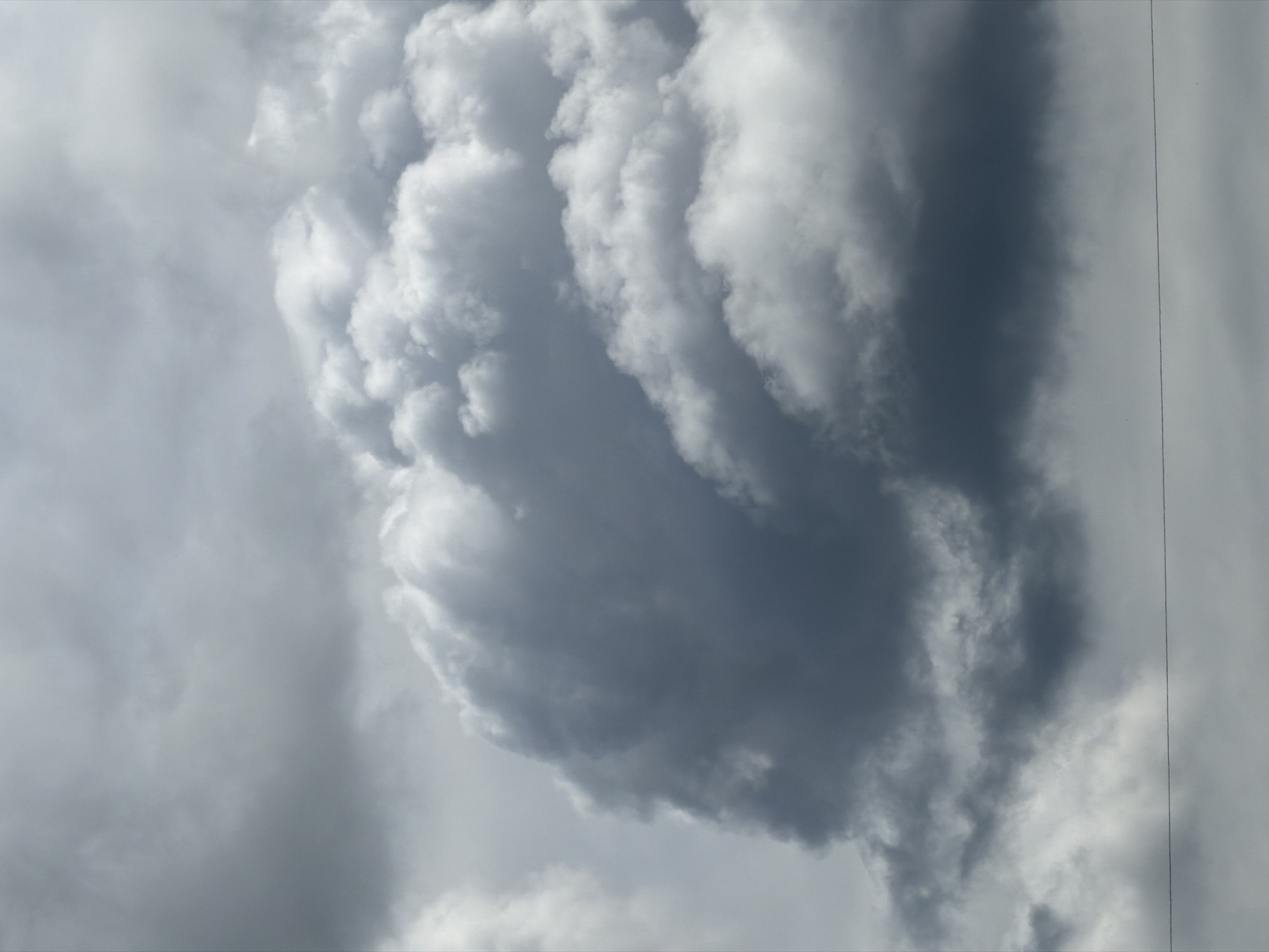 The image is a low-angle shot of a cloudy sky. The clouds are predominantly grey and white, with varying textures and densities. A prominent cloud formation in the center of the frame appears billowy and somewhat turbulent. A thin, vertical line is visible on the right side of the image, possibly a wire or scratch on the lens. There are no people or objects other than clouds in the frame. The environment is limited to the sky and clouds, offering no context regarding a specific location in Esch-sur-Alzette, Luxembourg.