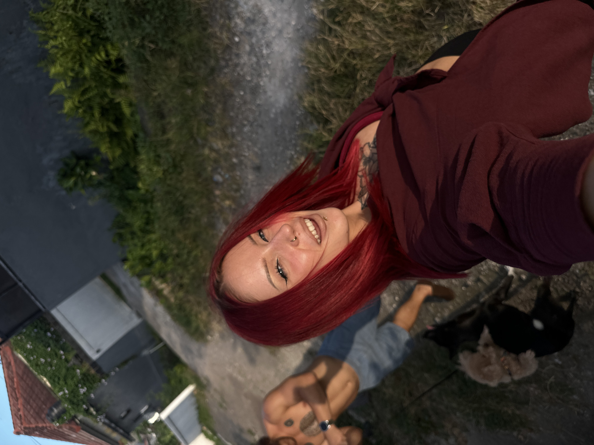 The image captures a selfie-style view with a young woman, positioned towards the top, smiling at the camera. She has red hair and facial piercings, and is wearing a maroon top. Visible tattoos are present on her neck and chest.
Below her, a man with visible chest tattoos wears light blue shorts. Two dogs are present, a black one and a smaller, light-colored one.
The setting appears to be outdoors. In the background, greenery, paved surfaces, and parts of buildings suggest an urban environment. Based on the location in North Kuta, Indonesia, the scene is probably a street or sidewalk with residential or commercial properties nearby.
