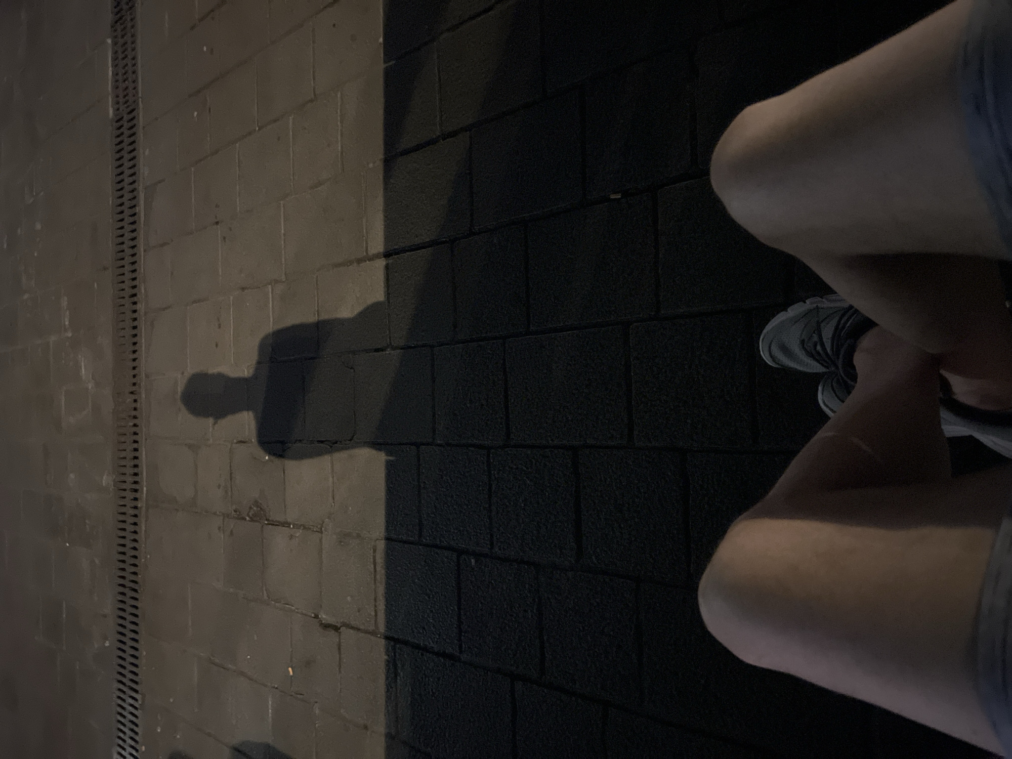 The image shows a first-person perspective of a person sitting on a tiled sidewalk at night. The person's legs and feet are in the foreground, with one leg crossed over the other. The person is wearing shorts and athletic shoes. In the background, a shadow of the person is cast on the sidewalk. The sidewalk is made of dark gray rectangular tiles, and a metal grate runs along the edge of the sidewalk. The scene appears to be in an outdoor urban environment.