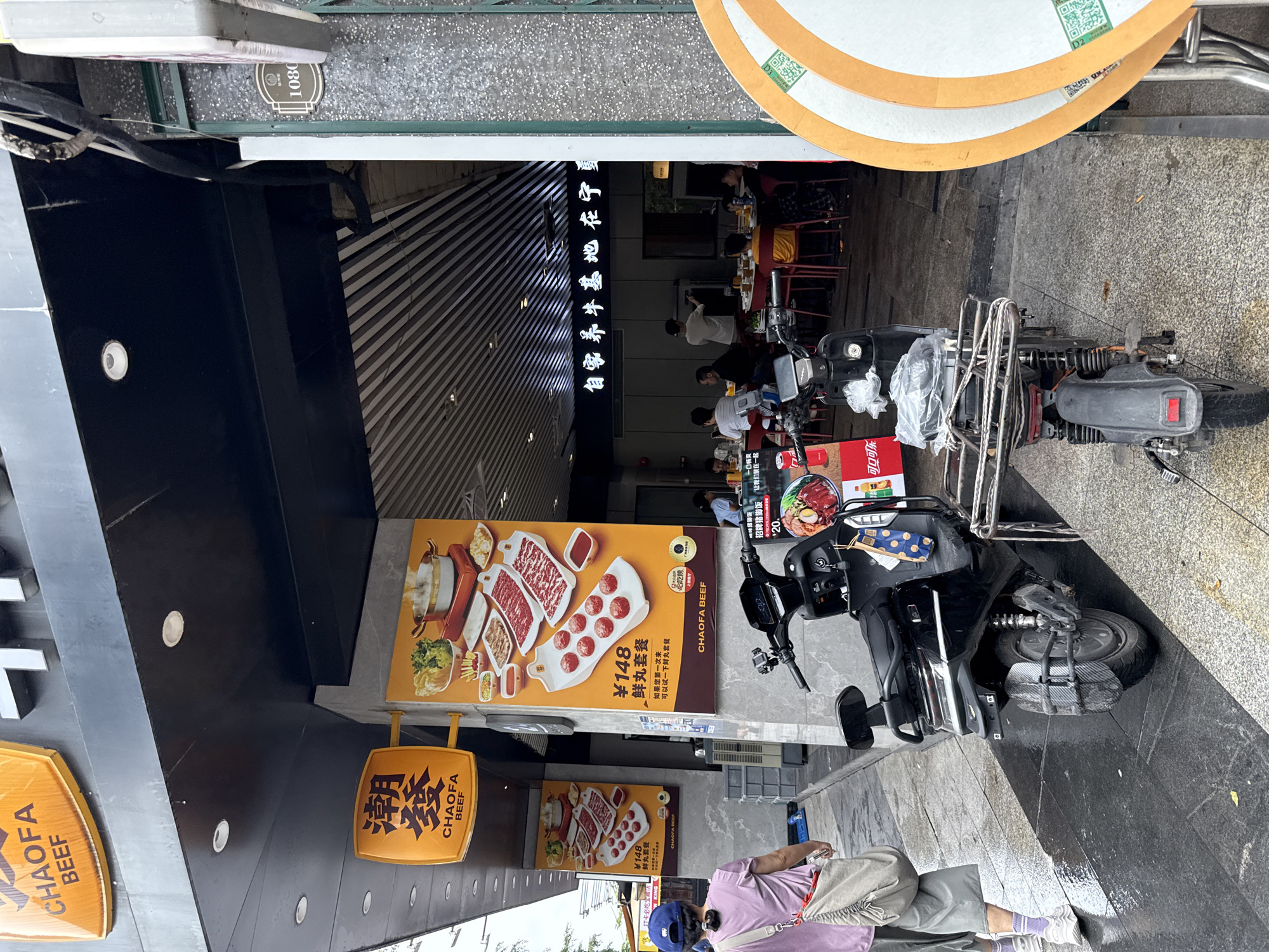 The image depicts the exterior of a restaurant in Haizhu District, China. A woman wearing a purple shirt and a blue cap walks away from the camera on a wet sidewalk. Two electric scooters are parked in front of the restaurant entrance. The restaurant has large posters advertising beef dishes, with Chinese characters and "CHAOFA BEEF" branding. Inside the restaurant, several patrons are seated at tables. The architecture of the building includes a black overhang with strip lighting. A building number "1080" is visible on a plaque to the right of the entrance.