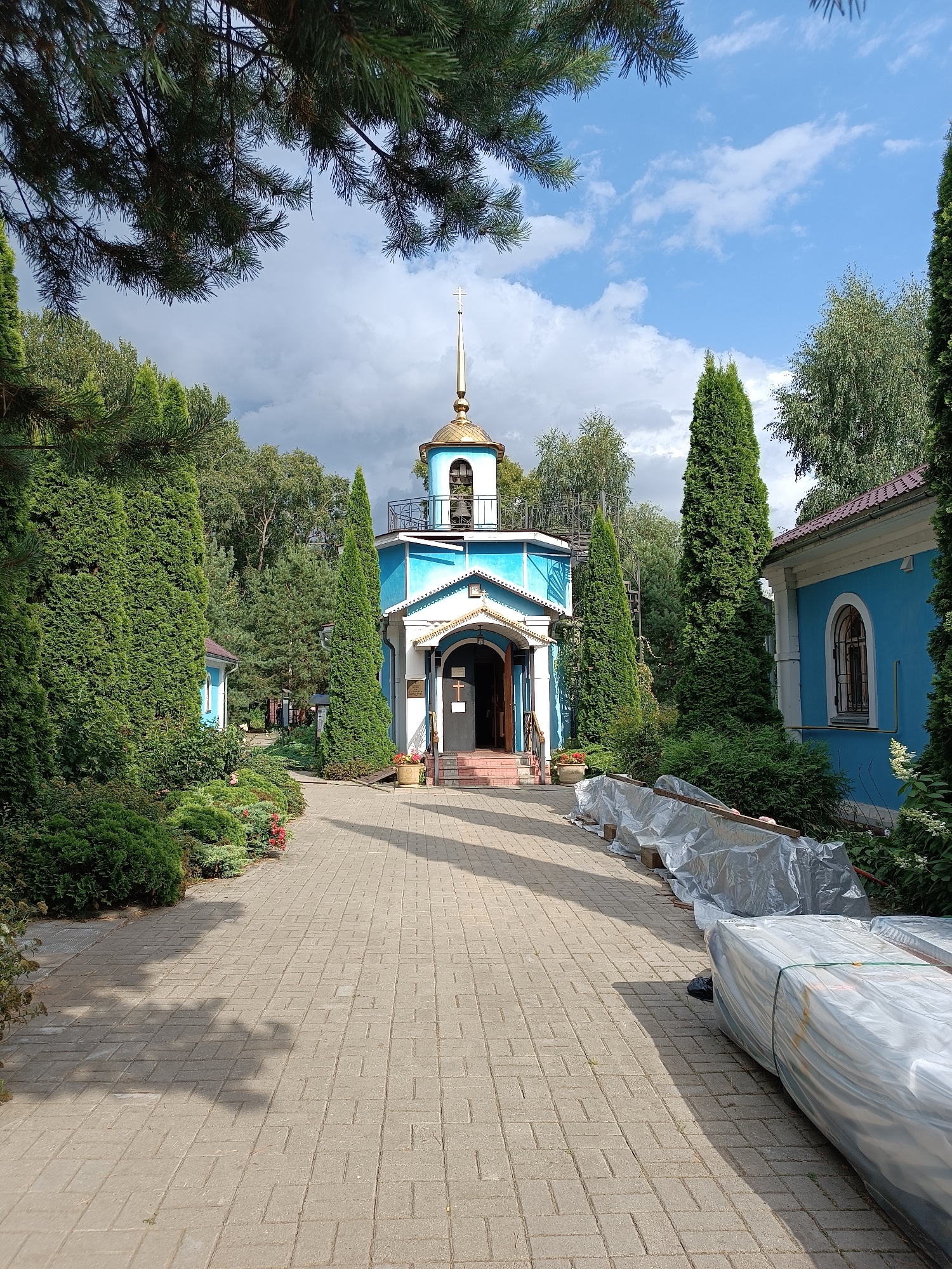 The image shows a pathway leading to a blue and white church with a golden dome and cross. The pathway is paved with rectangular bricks and is lined with trimmed green hedges and cypress trees. In the background, there are other trees and a portion of another blue building. The sky is partly cloudy with some sun. There are some materials covered in plastic sheeting on the right side of the pathway. The church appears to be the Church of the Transfiguration in Bykovo, Russia.