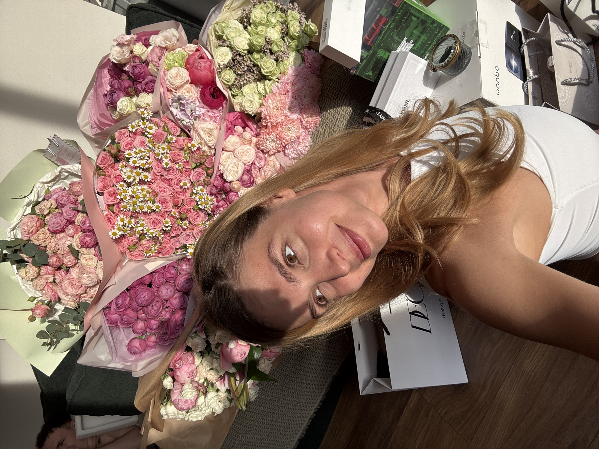 The image shows a woman with long, light brown hair, wearing a white top, lying on the floor next to multiple bouquets of flowers. The flowers are various colors, including pink, purple, white, and green. Next to the woman and flowers are several boxes and shopping bags, with a "deo.ge" logo visible on one bag. Objects such as sunglasses and a small decorative object are also present. The scene appears to be indoors, with a wooden floor and a sofa visible in the background. The location is indicated as the city of Unknown, Unknown.